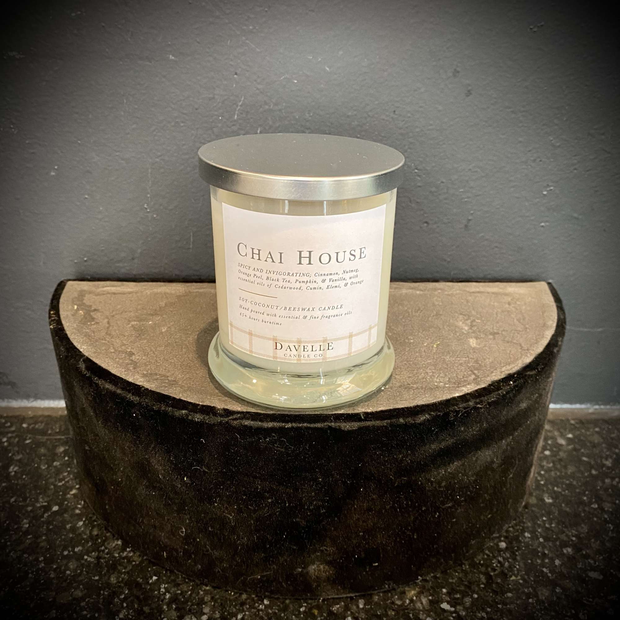 A glass jar candle with a silver lid sits on a dark velvet stand.
