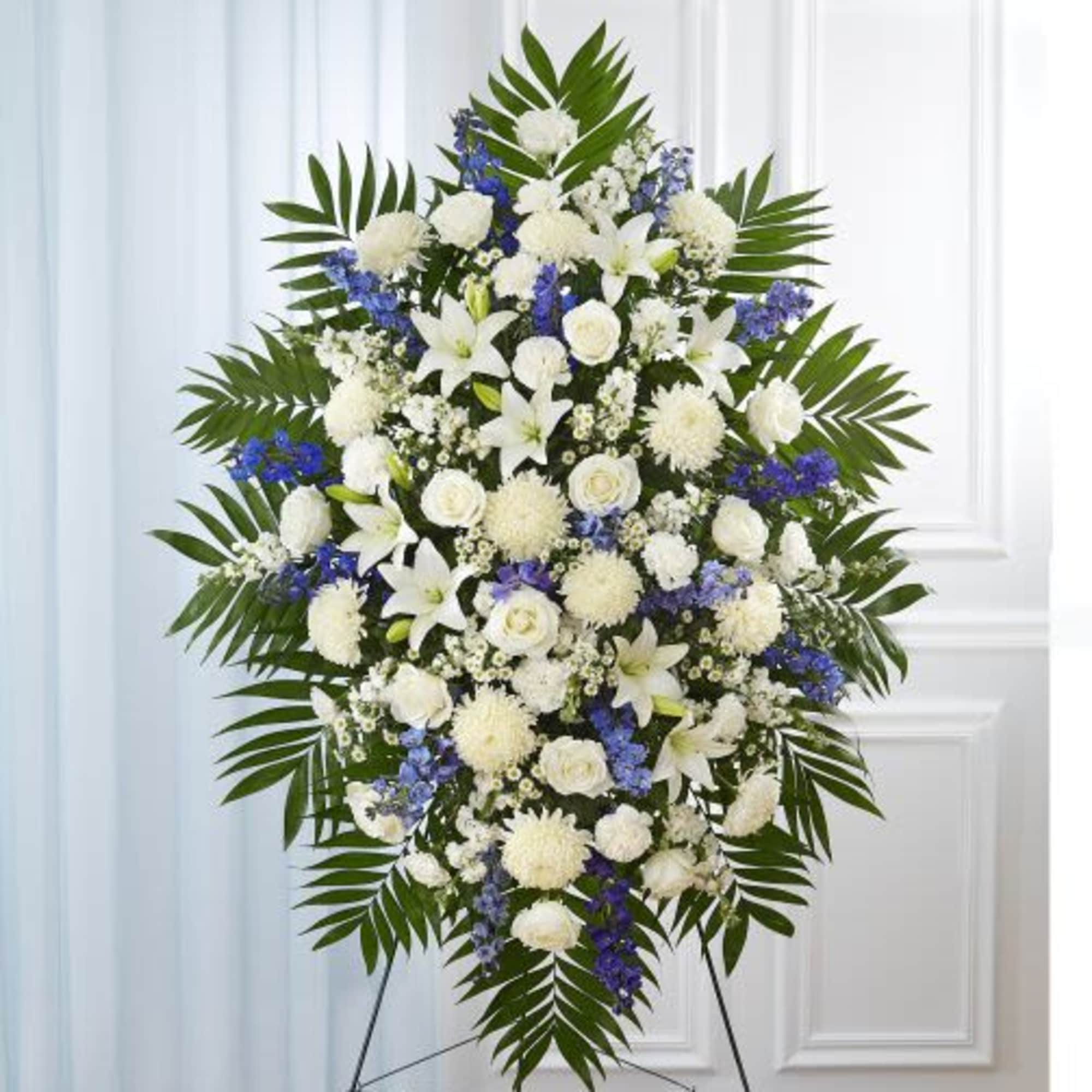 Tall standing spray of white roses, lilies, mums and blue flowers with palm fronds on an easel