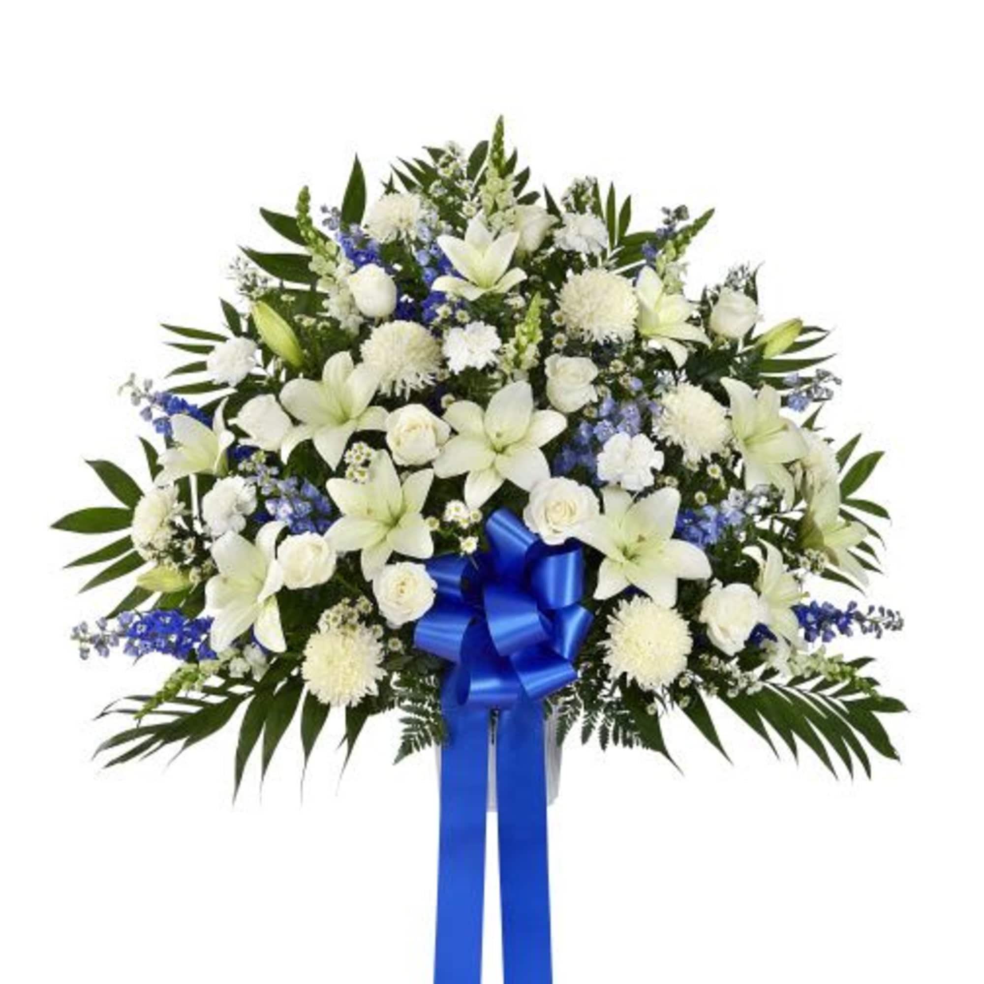 Standing spray of white lilies, roses, and mums with blue flowers and a large blue ribbon bow on an easel