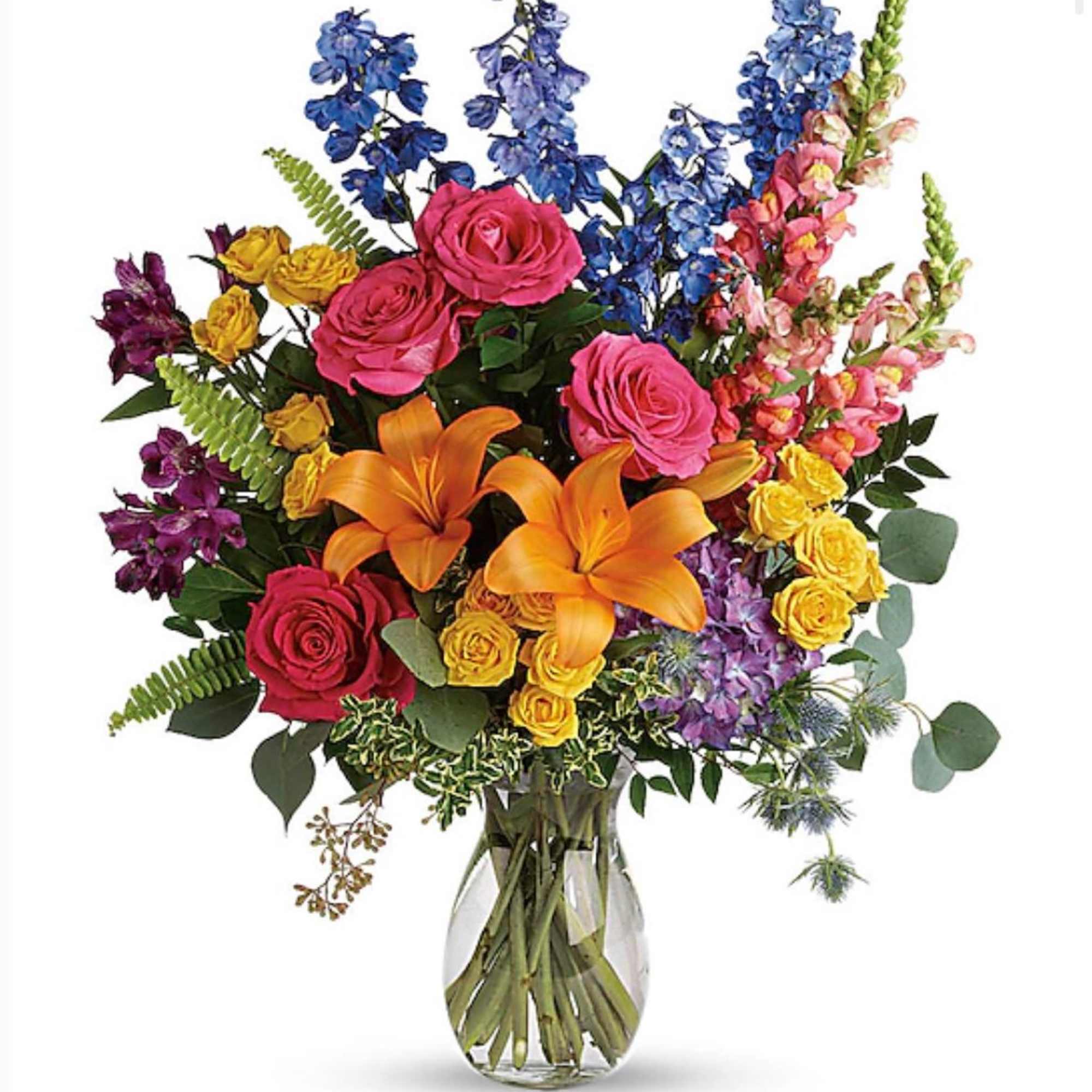 Tall mixed bouquet with orange lilies, pink and yellow roses, and blue delphinium in a clear glass vase.