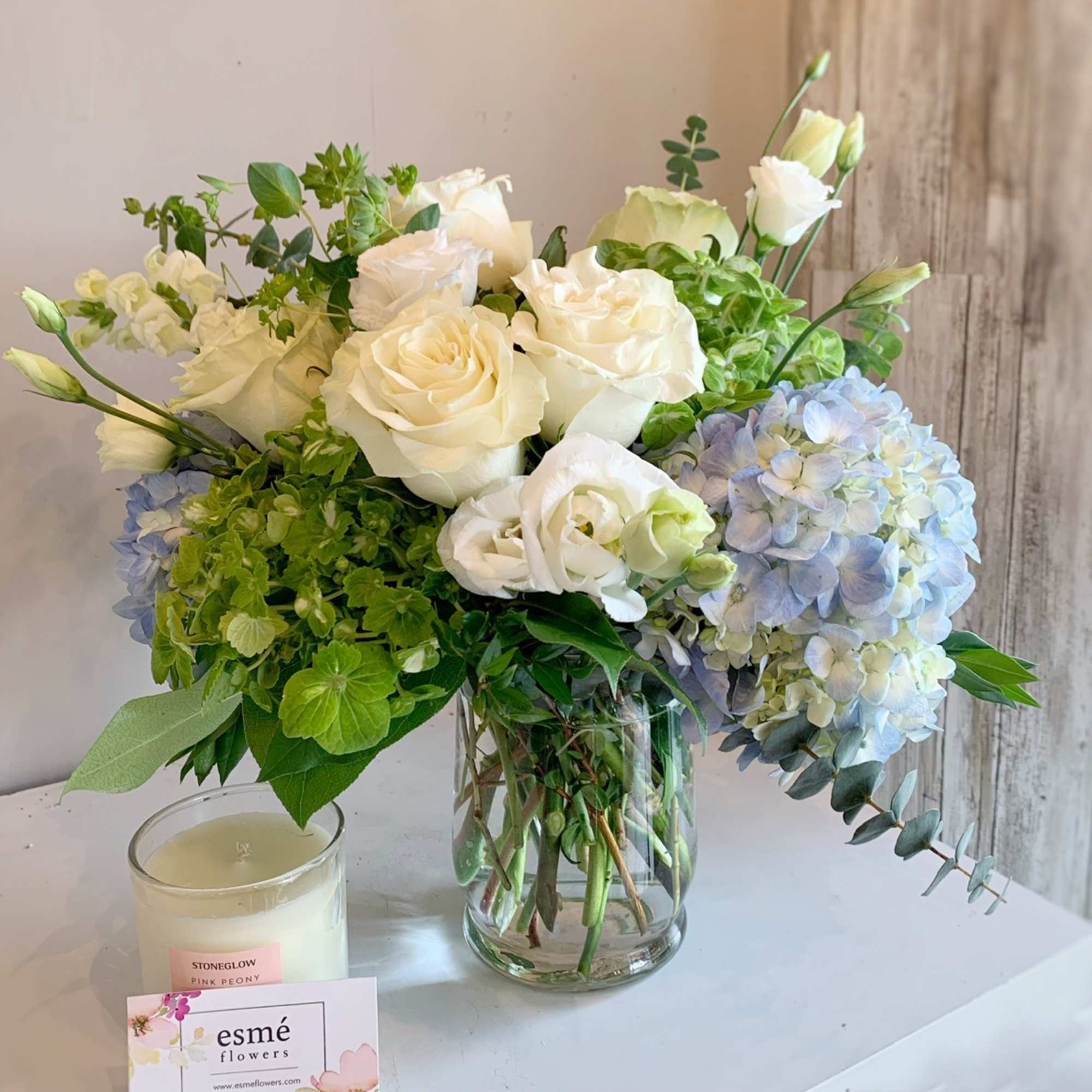 White roses and blue hydrangeas in a glass vase with a candle beside it.