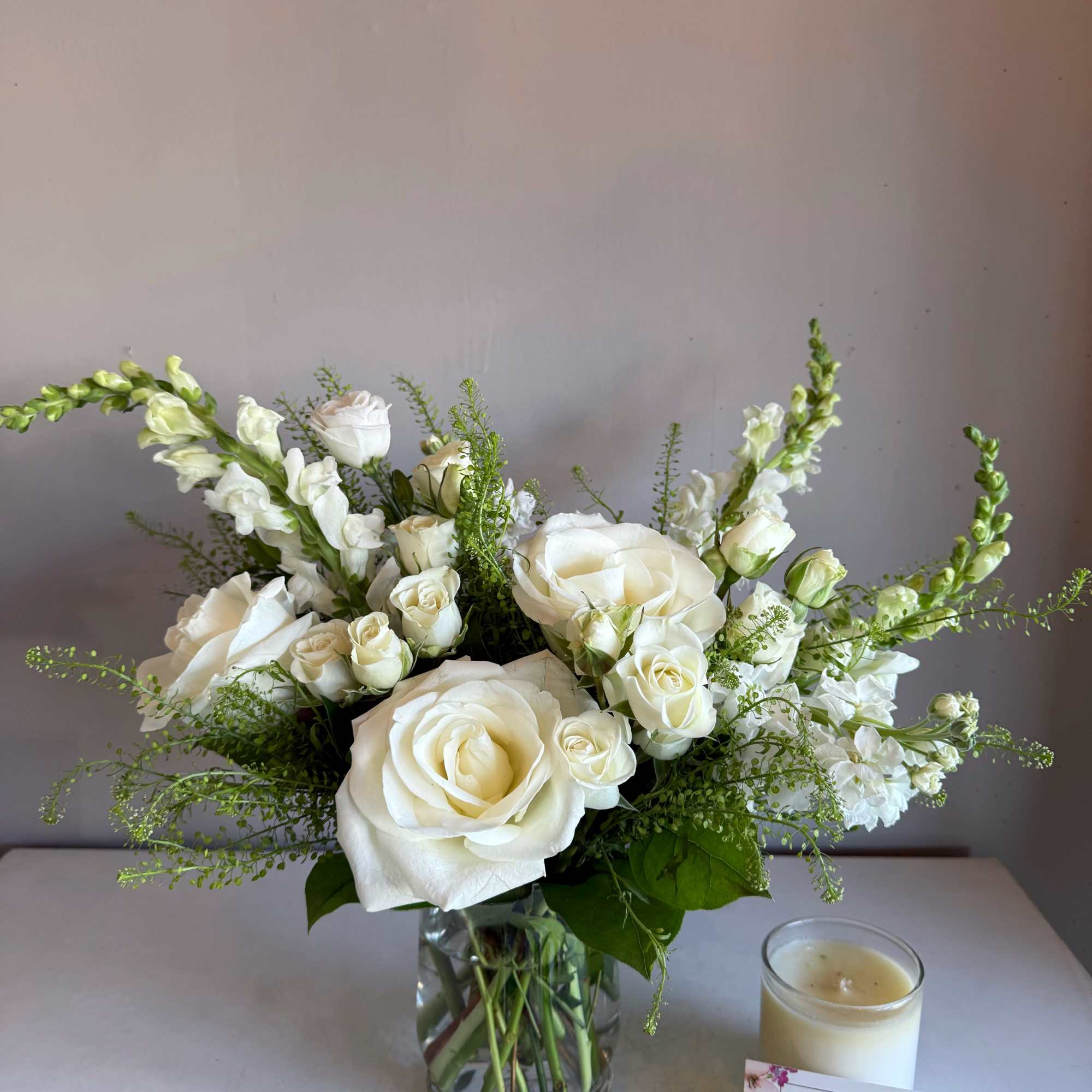 White roses and snapdragons arranged in a glass vase beside a candle.