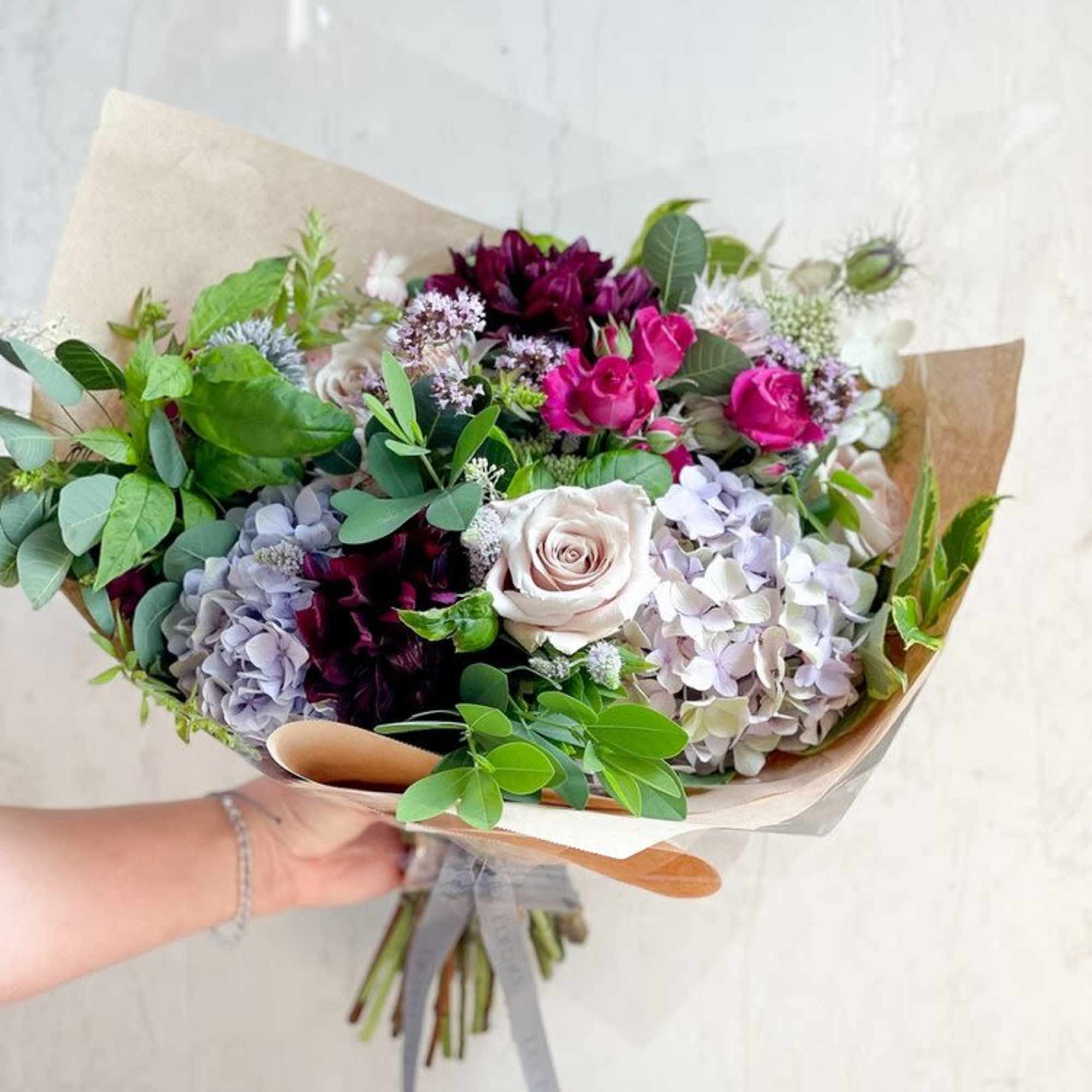 Hand-tied bouquet of roses, hydrangeas, and dark blooms wrapped in paper
