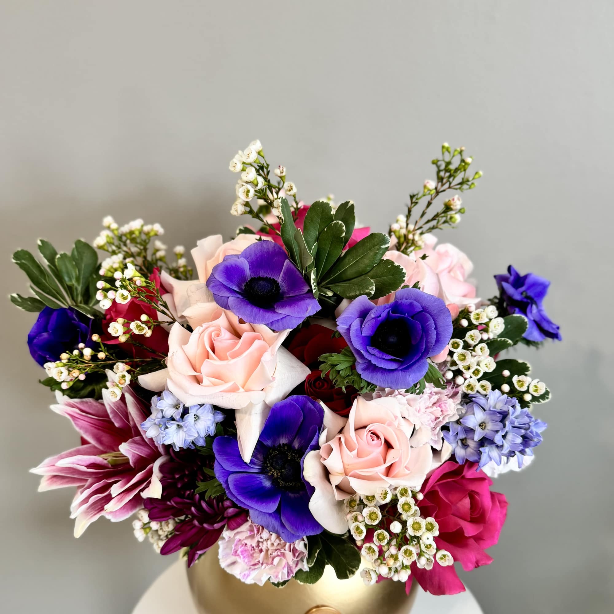 Low arrangement of purple anemones, pink roses, and mixed blooms in a matte gold bowl vase