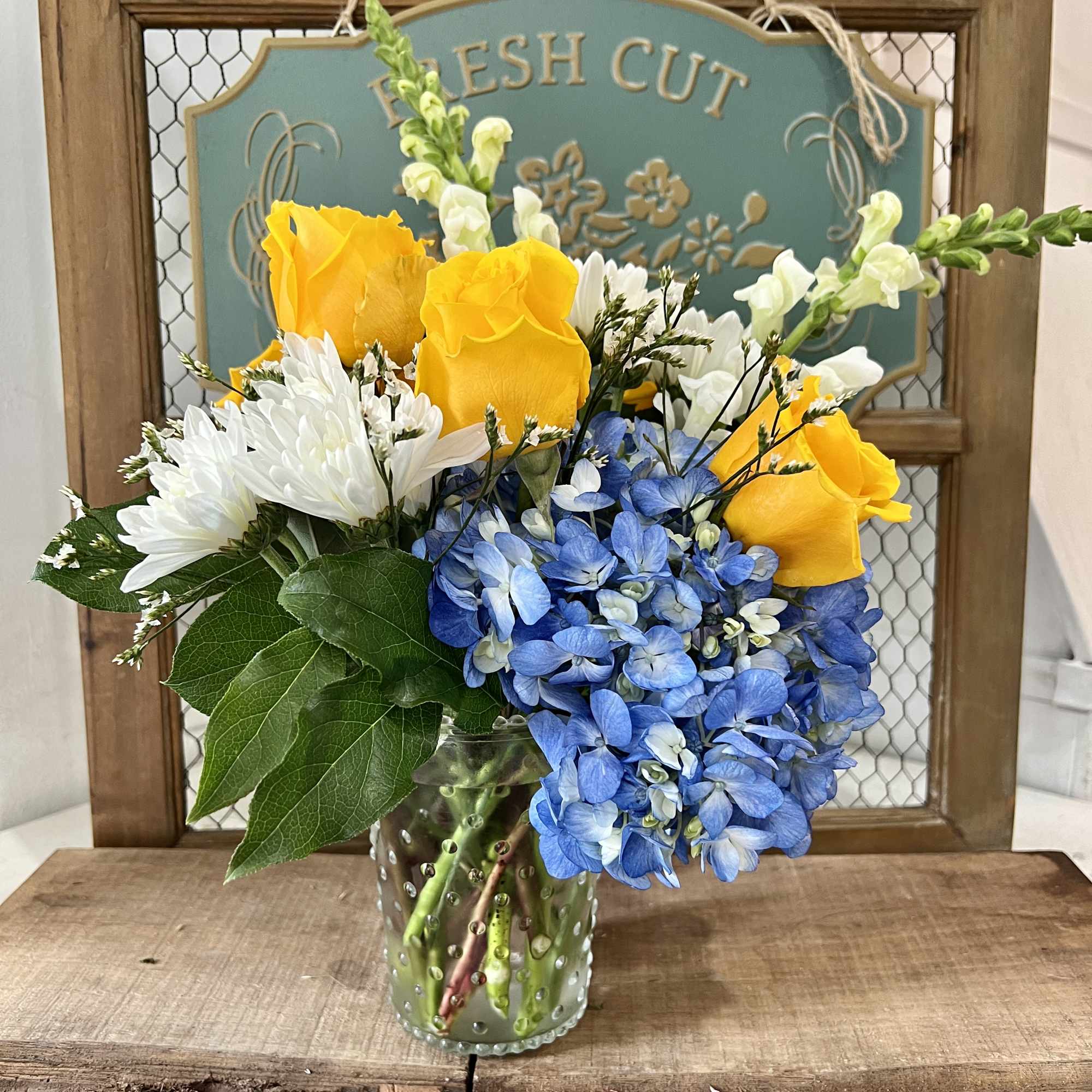 Yellow roses, blue hydrangeas, and white daisies in a glass vase