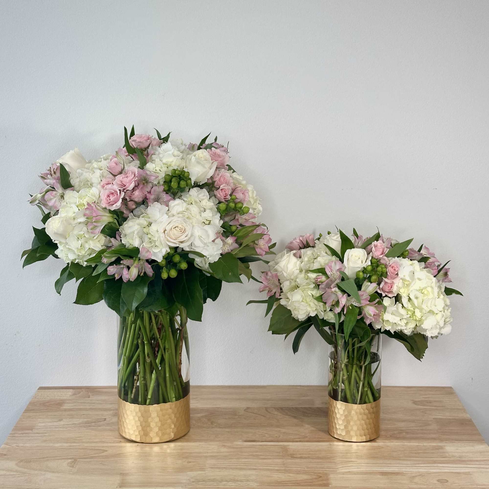 Two pink and white floral arrangements in gold vases on a wooden table