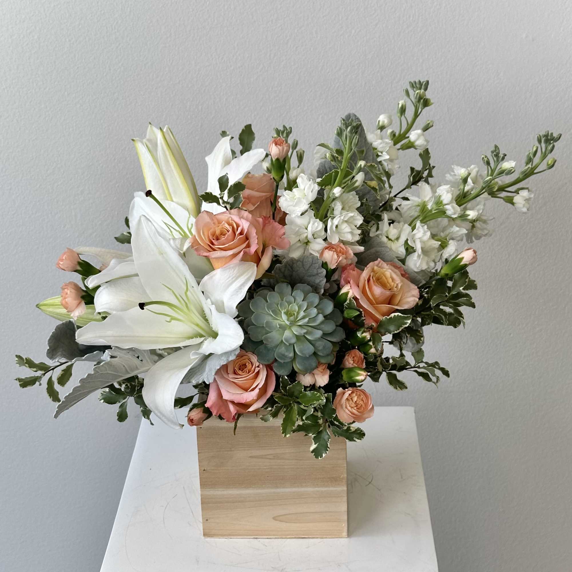 Peach roses, white lilies, stock, and a succulent arranged in a wooden cube container
