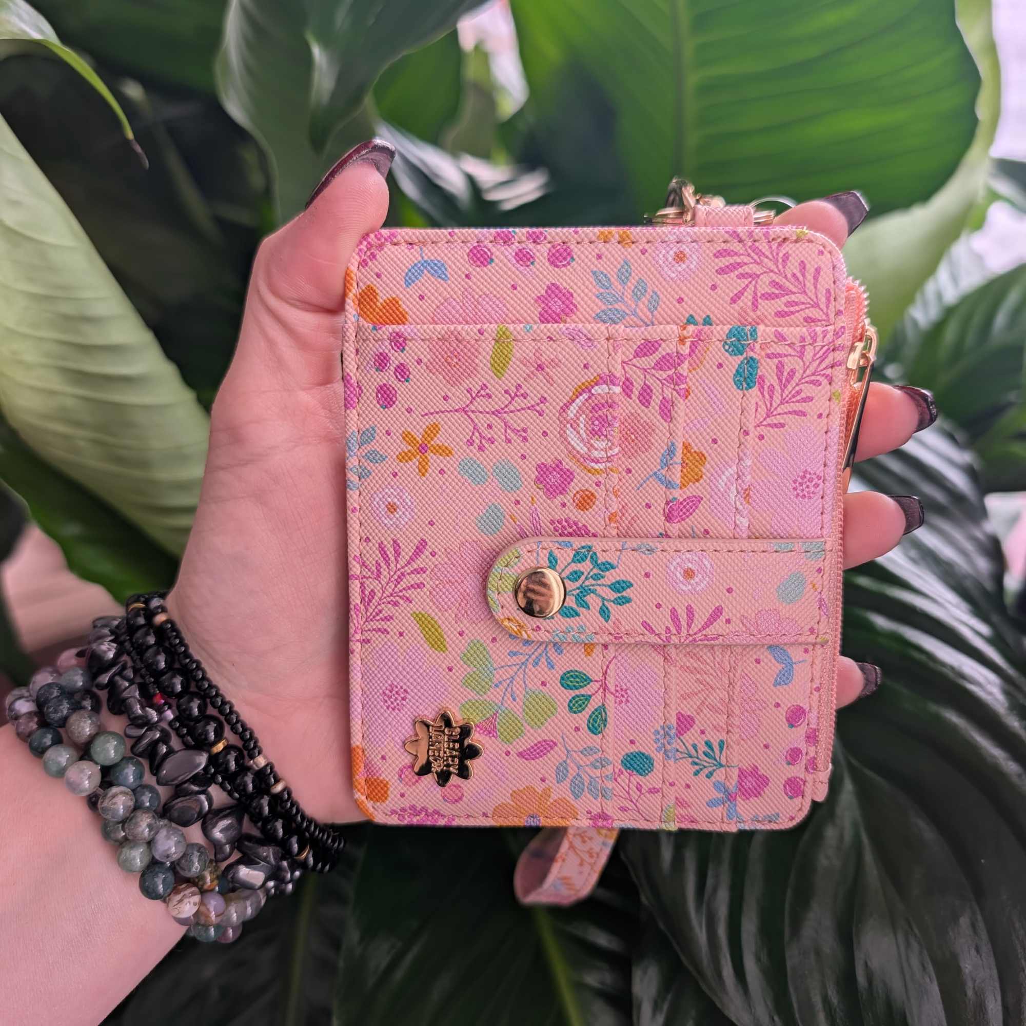 Hand holding a pink floral wallet against large green leaves