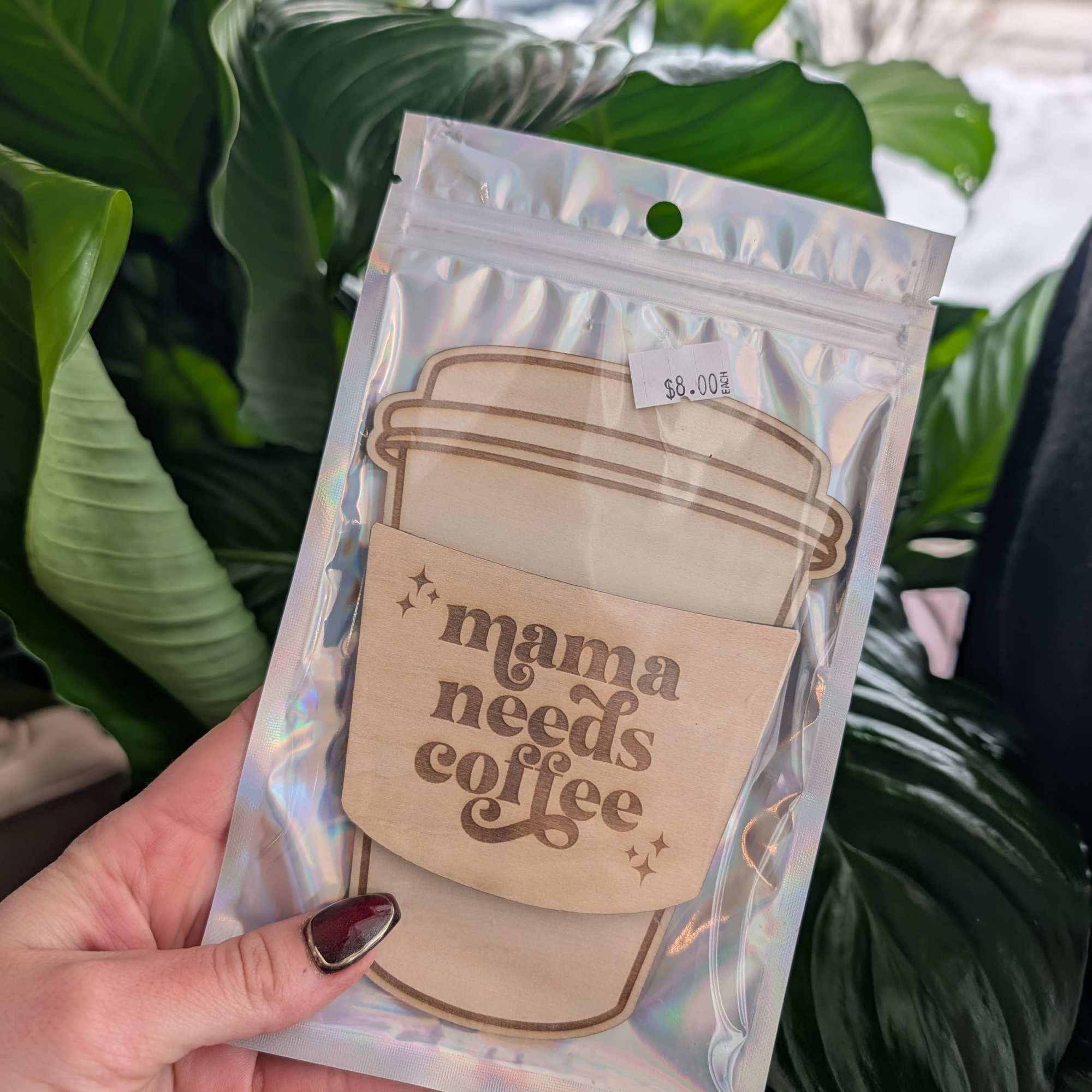 Wooden coffee-themed sign in a clear pouch held in front of green houseplants