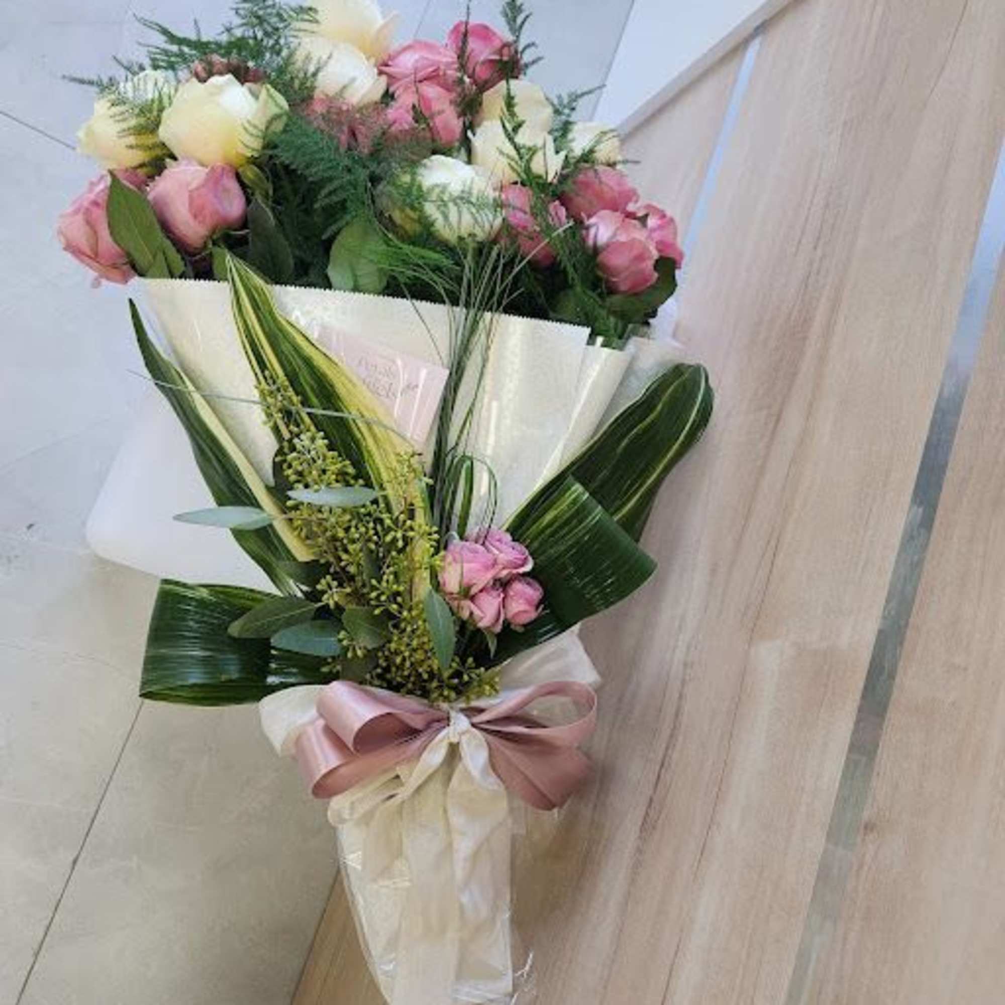 Hand-tied bouquet of pink and cream roses with greenery, wrapped in white paper and ribbon.