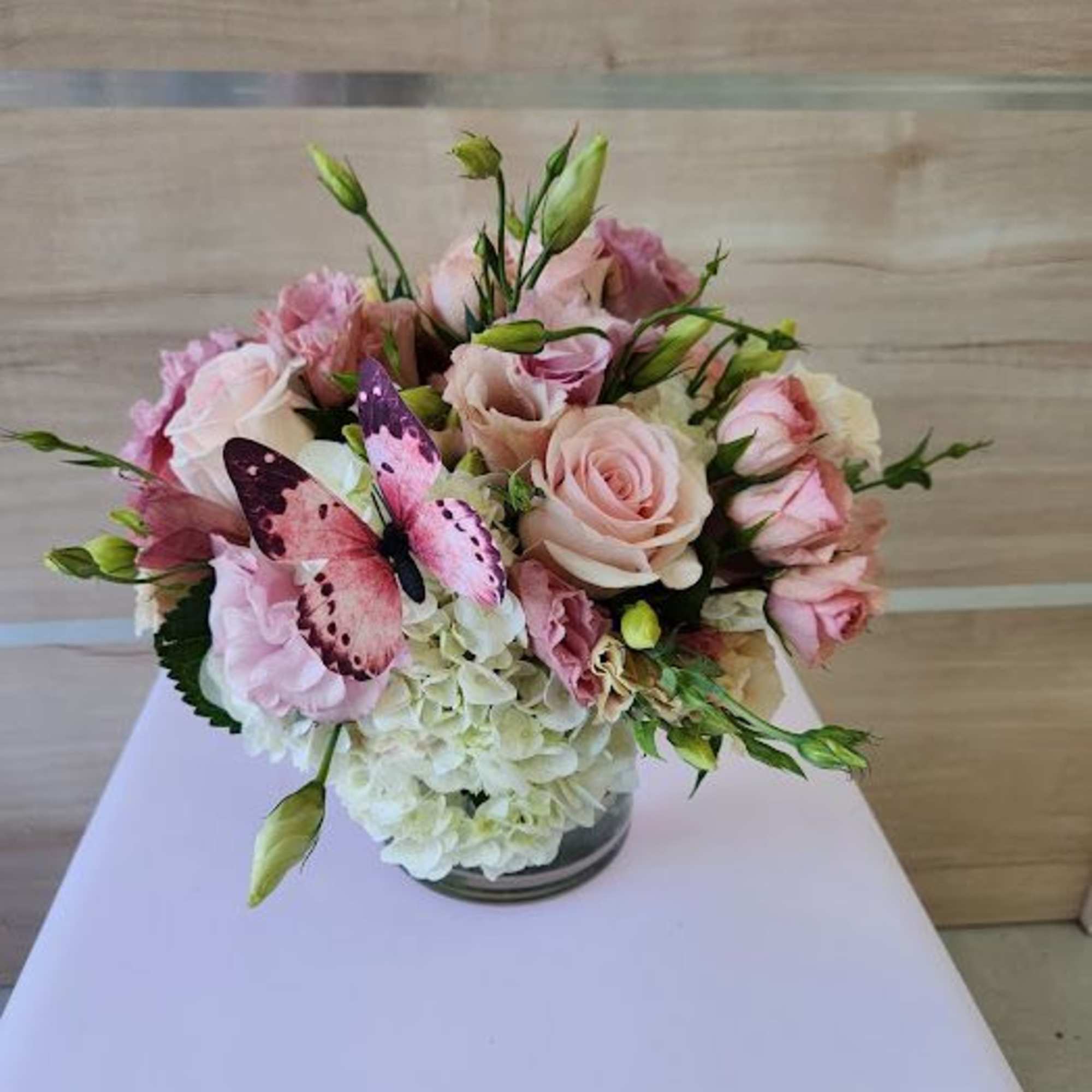 Low arrangement of blush pink roses and lisianthus with white hydrangeas in a glass vase and a pink butterfly accent