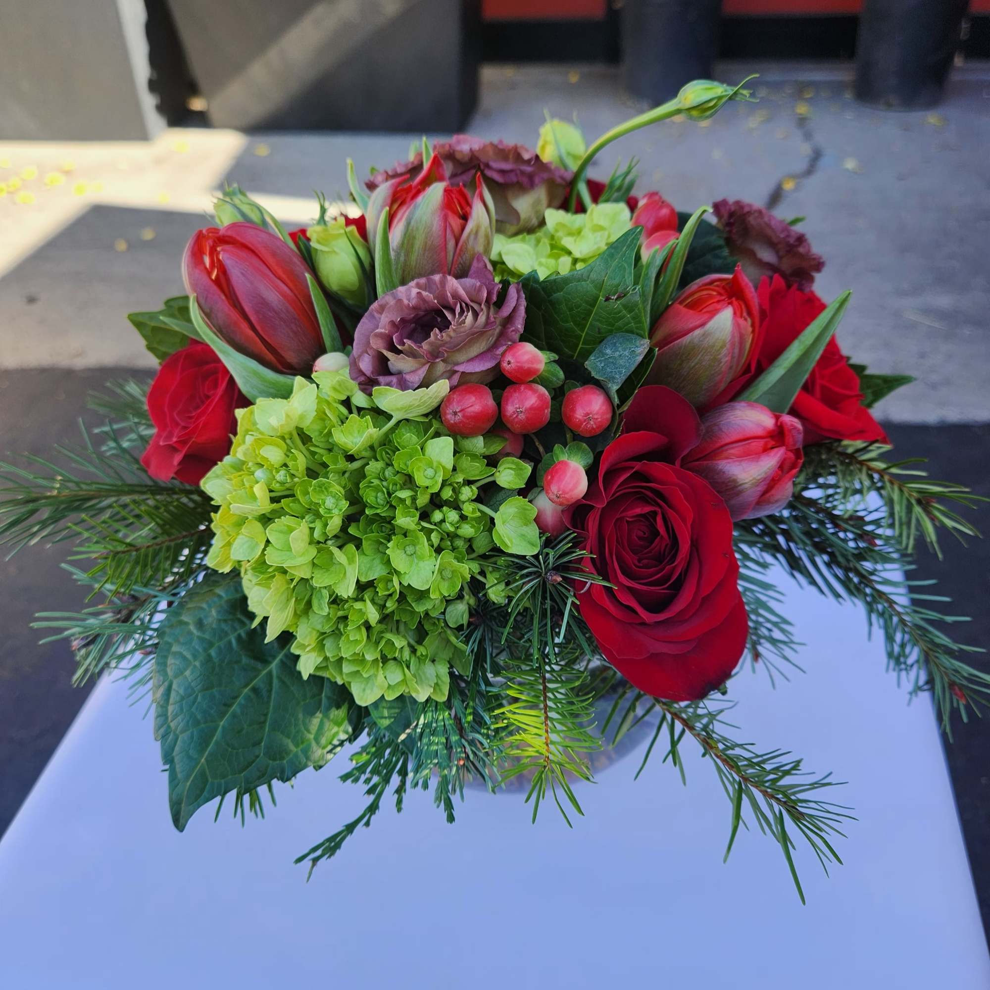 Short arrangement of red roses, tulips, green hydrangea, berries and lisianthus in a glass vase with evergreen sprigs.
