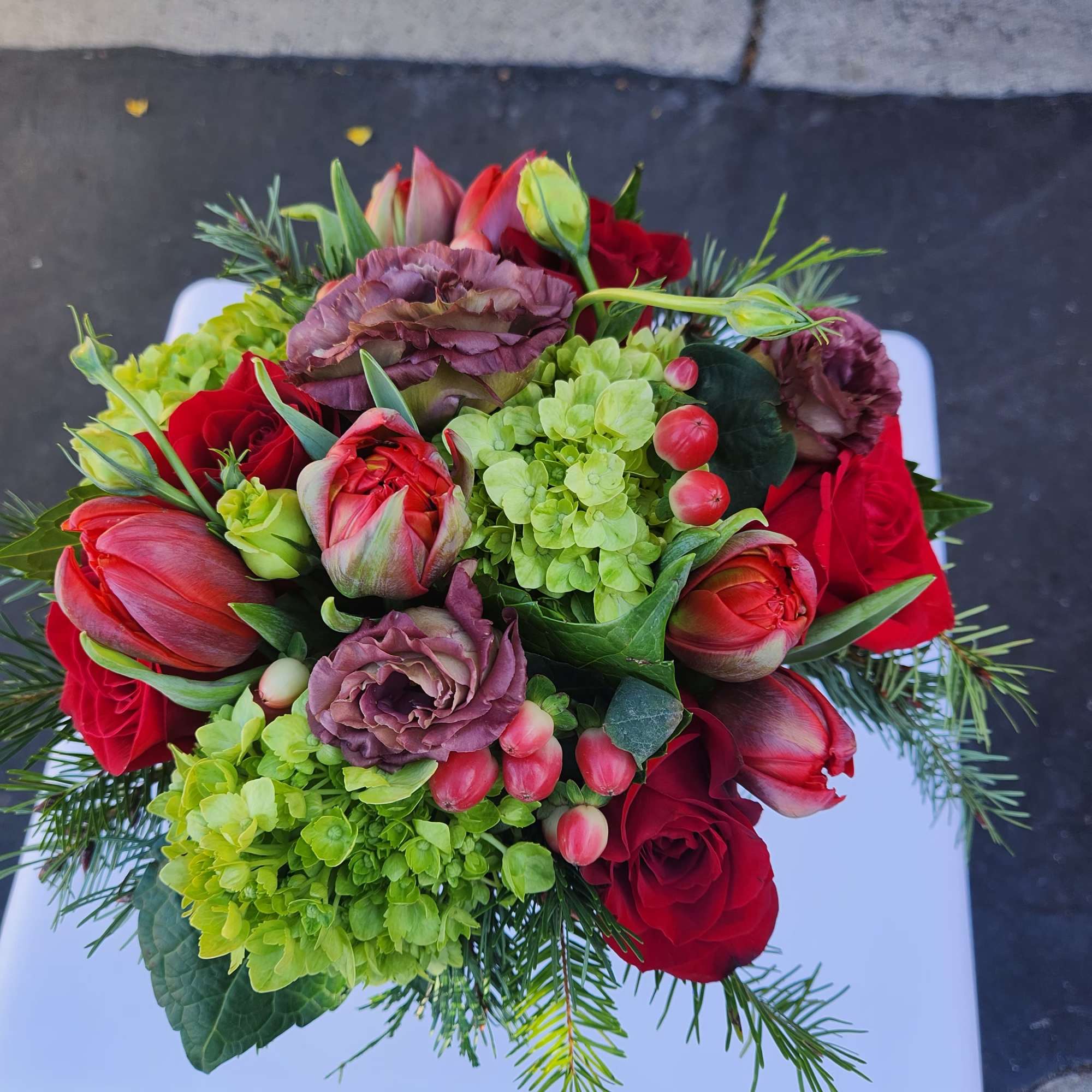 Compact arrangement of red roses and tulips with green hydrangea and mauve blooms on a white stand