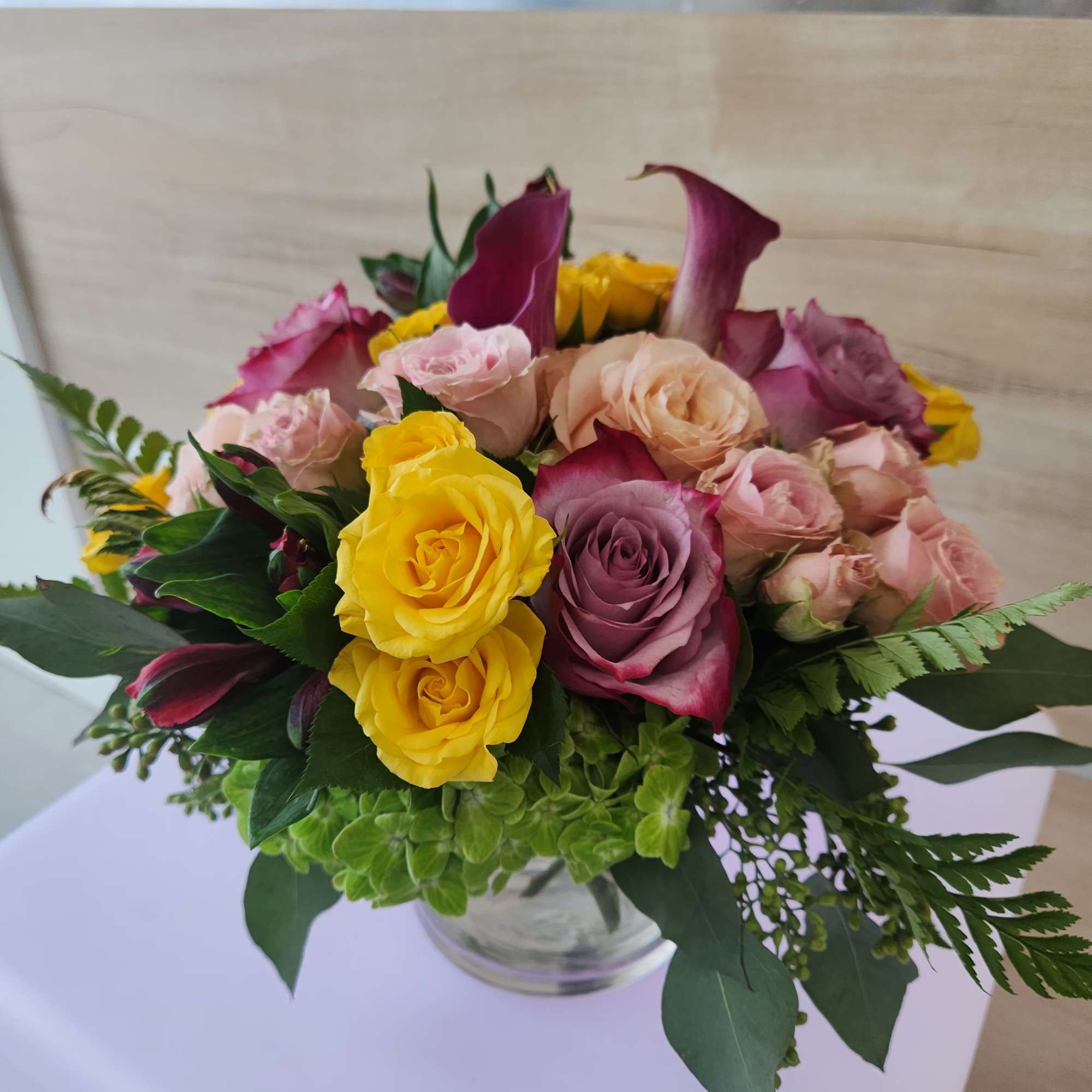 Round arrangement of yellow and pink roses with purple calla lilies in a clear glass vase