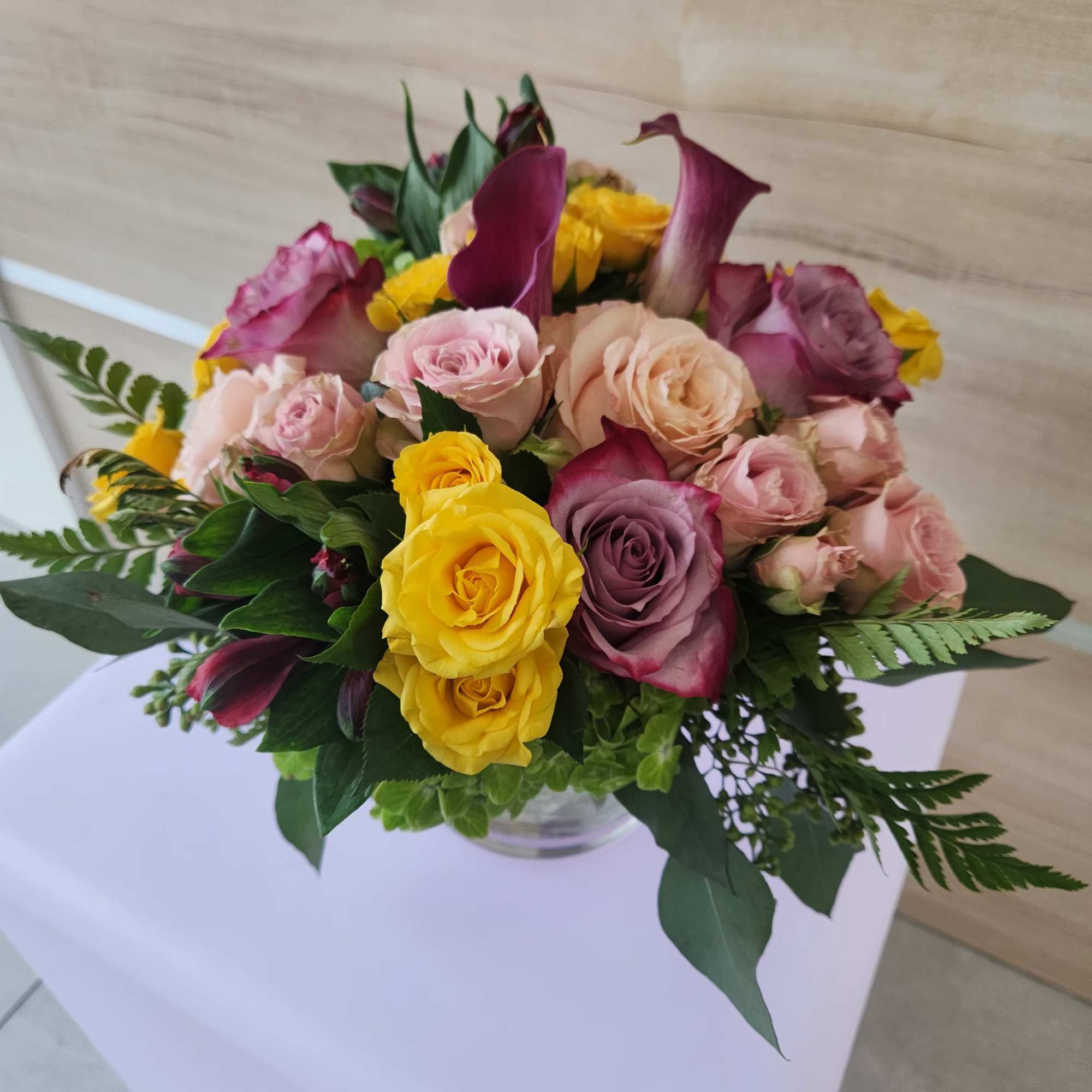 Low arrangement of yellow and pink roses with purple mini calla lilies in a clear glass vase