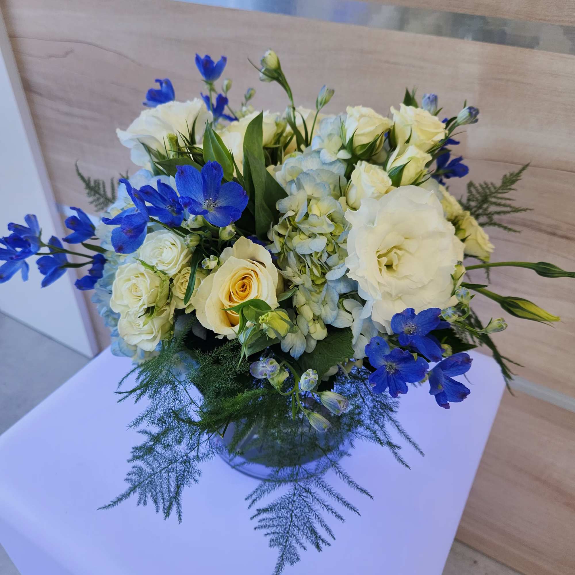 Low round arrangement of white and cream flowers with blue accents in a clear glass vase