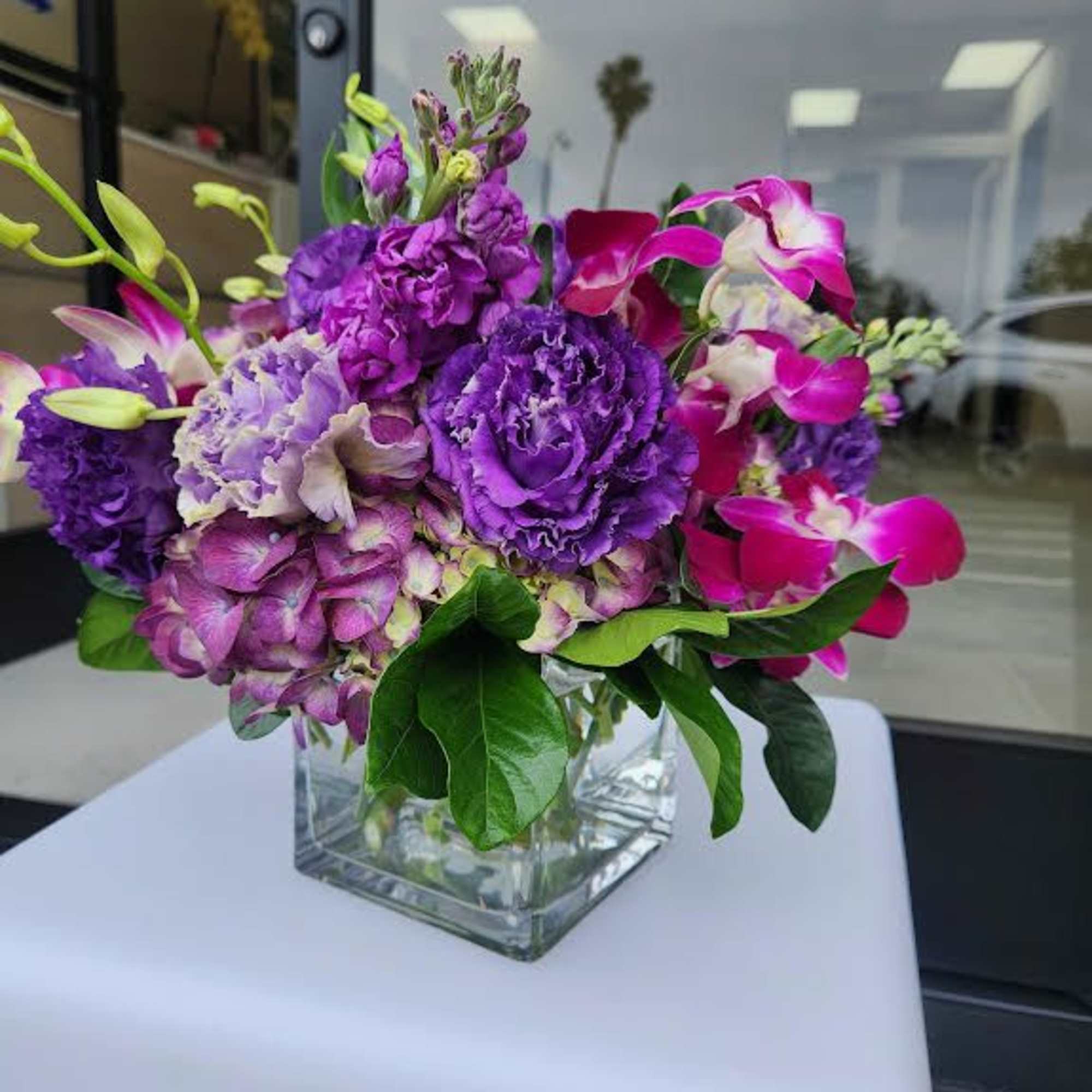 Compact arrangement of purple and pink flowers in a clear glass cube vase.