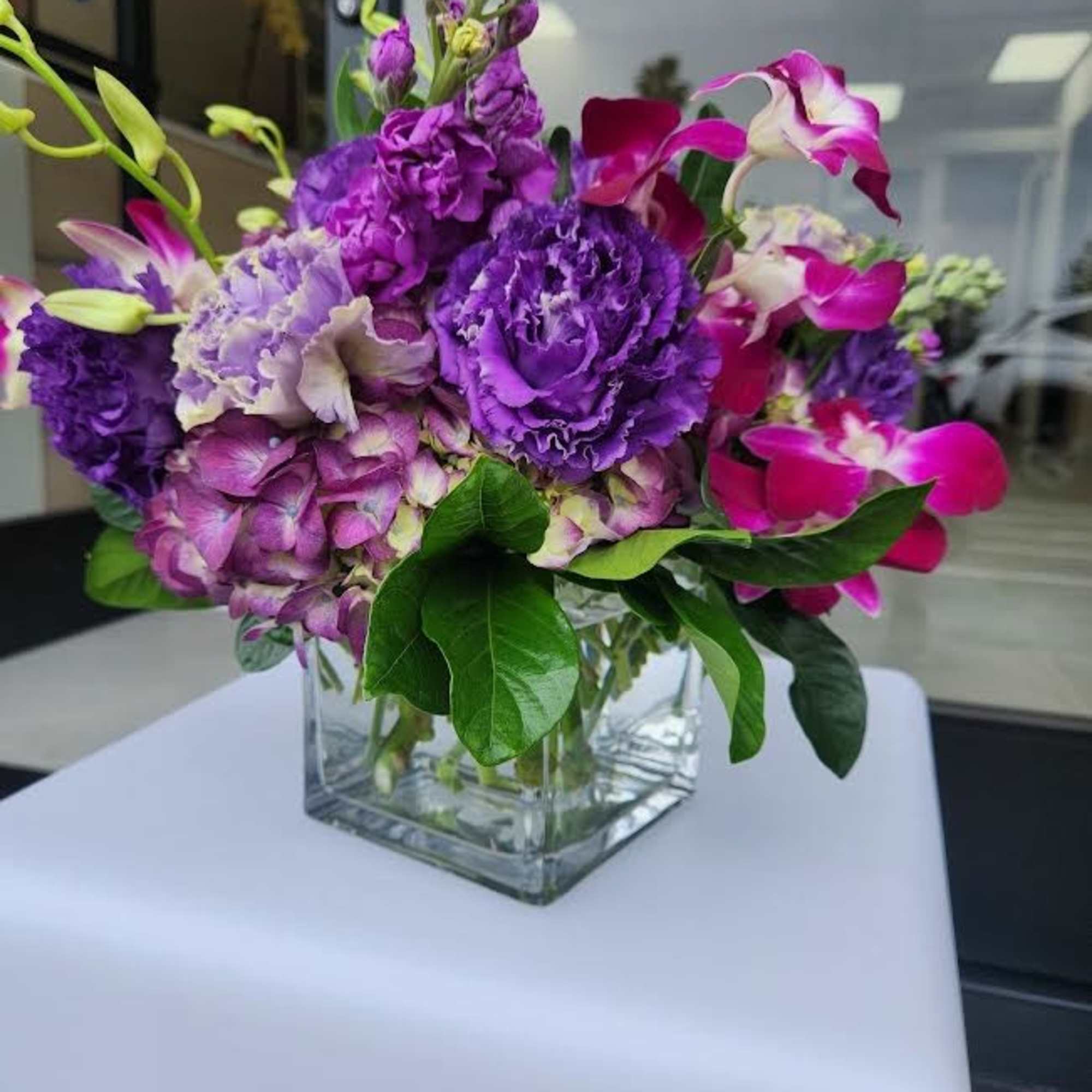 Compact purple and pink flower arrangement in a clear glass cube vase on a white stand.