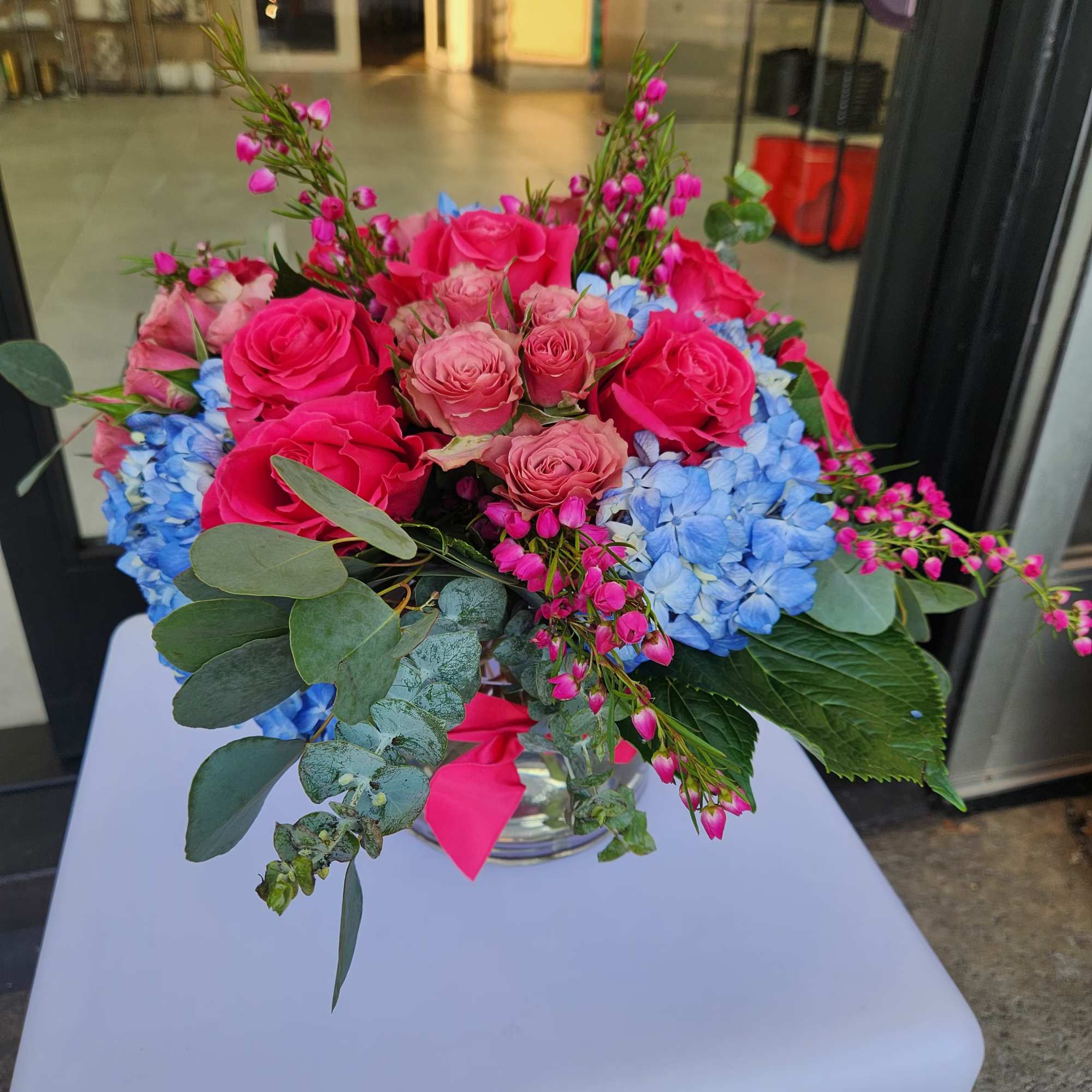 Low glass vase arrangement with hot pink roses and blue hydrangeas accented by small pink blossoms.