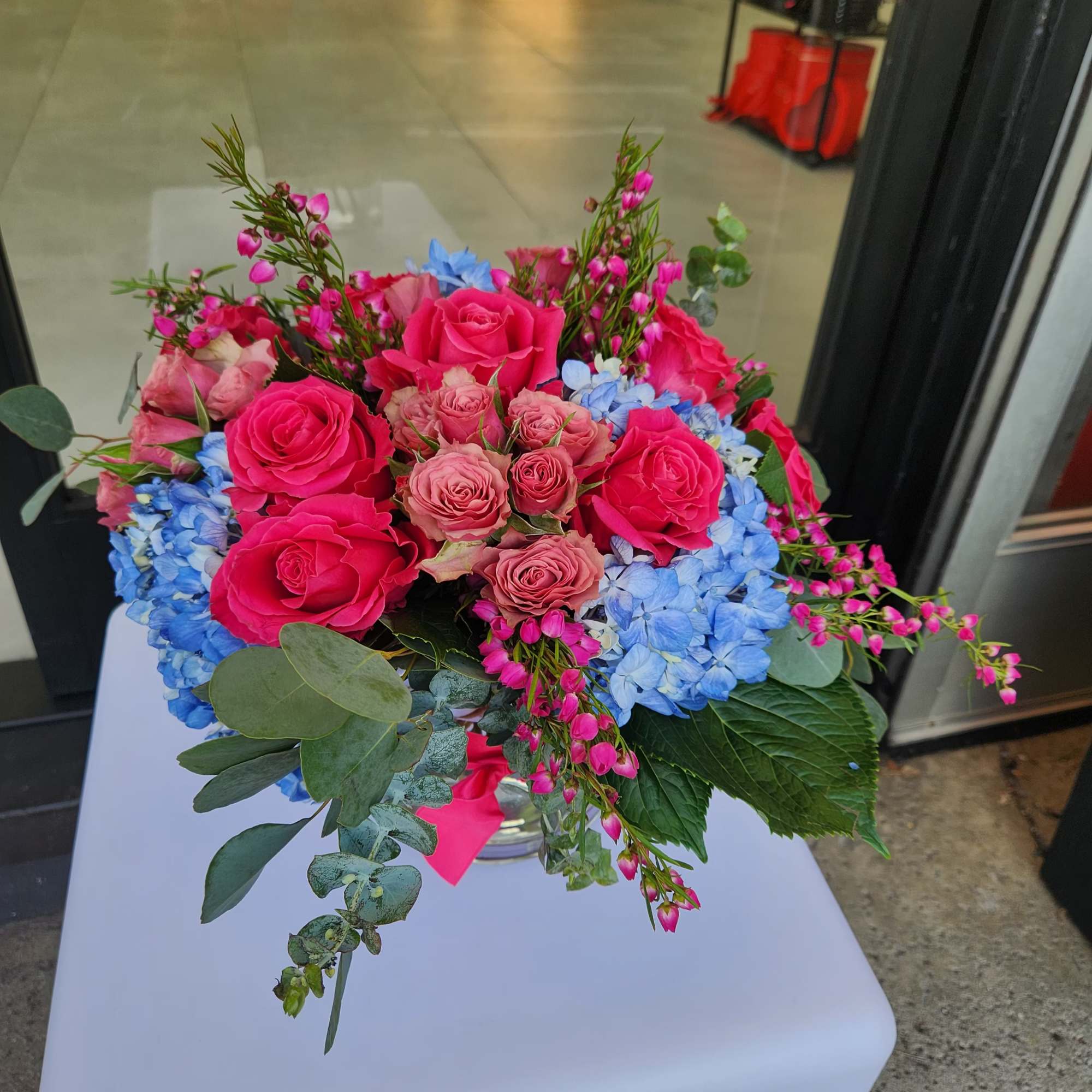 Round arrangement of hot pink roses, blush roses, and blue hydrangeas in a clear vase with greenery
