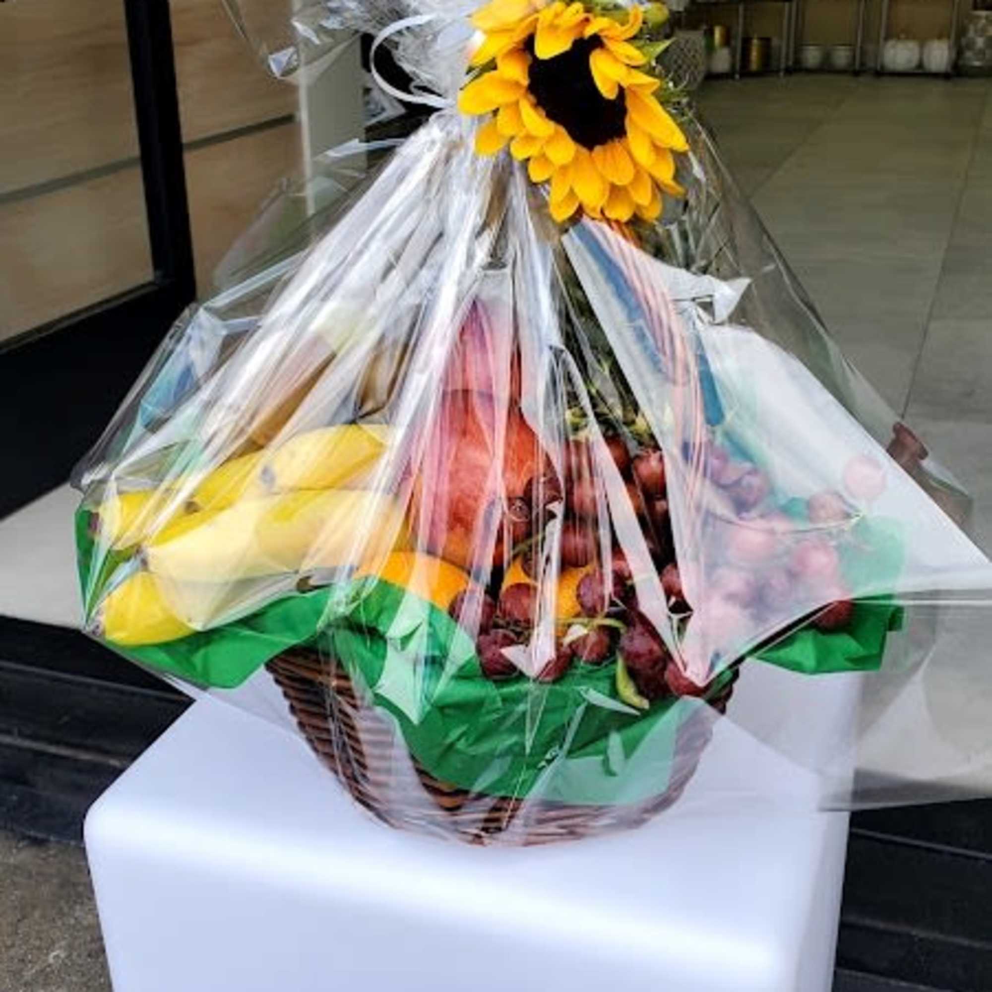 Gift basket of assorted fresh fruit wrapped in cellophane and topped with a single yellow sunflower.