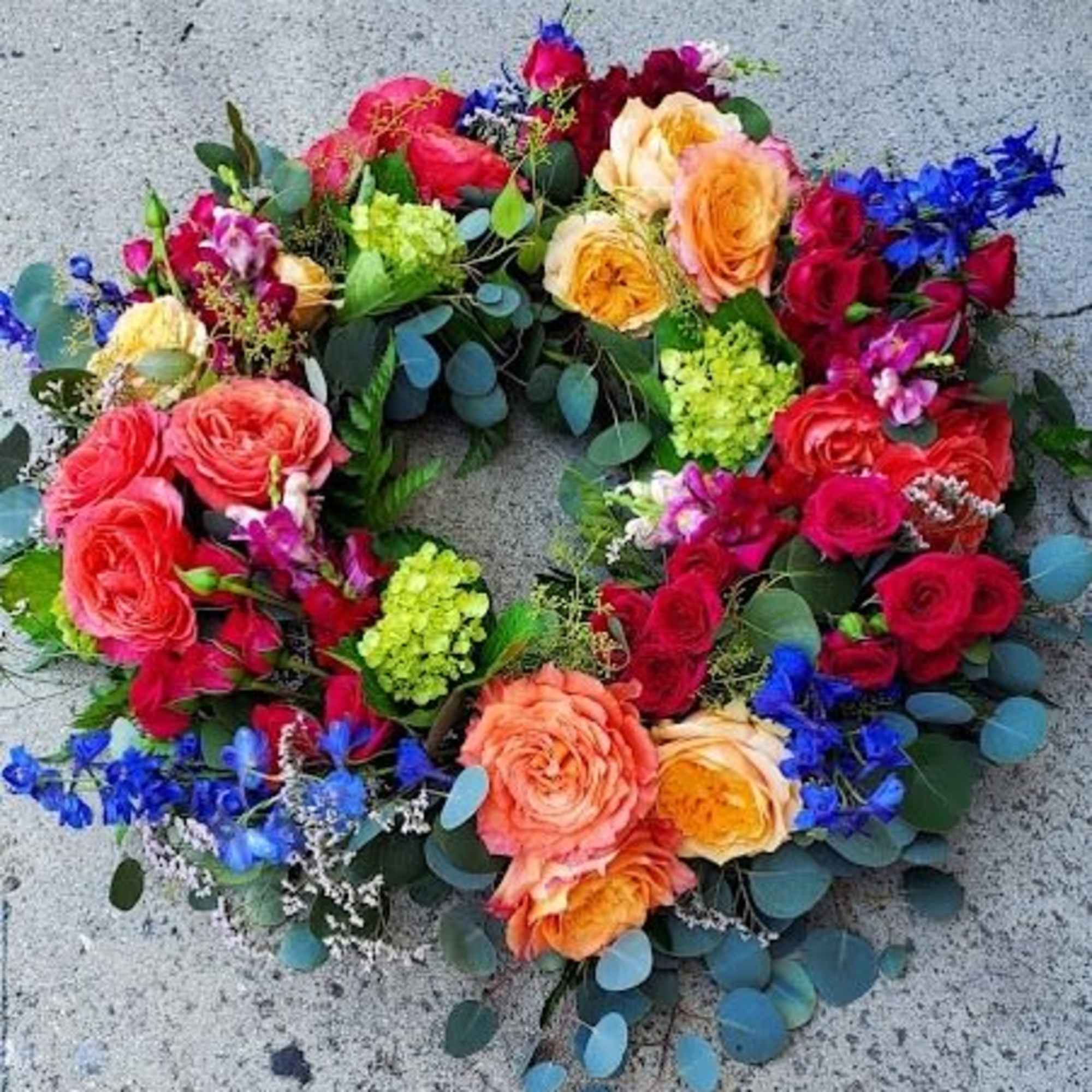 Bright circular wreath of roses, hydrangea, and blue delphinium on a concrete surface
