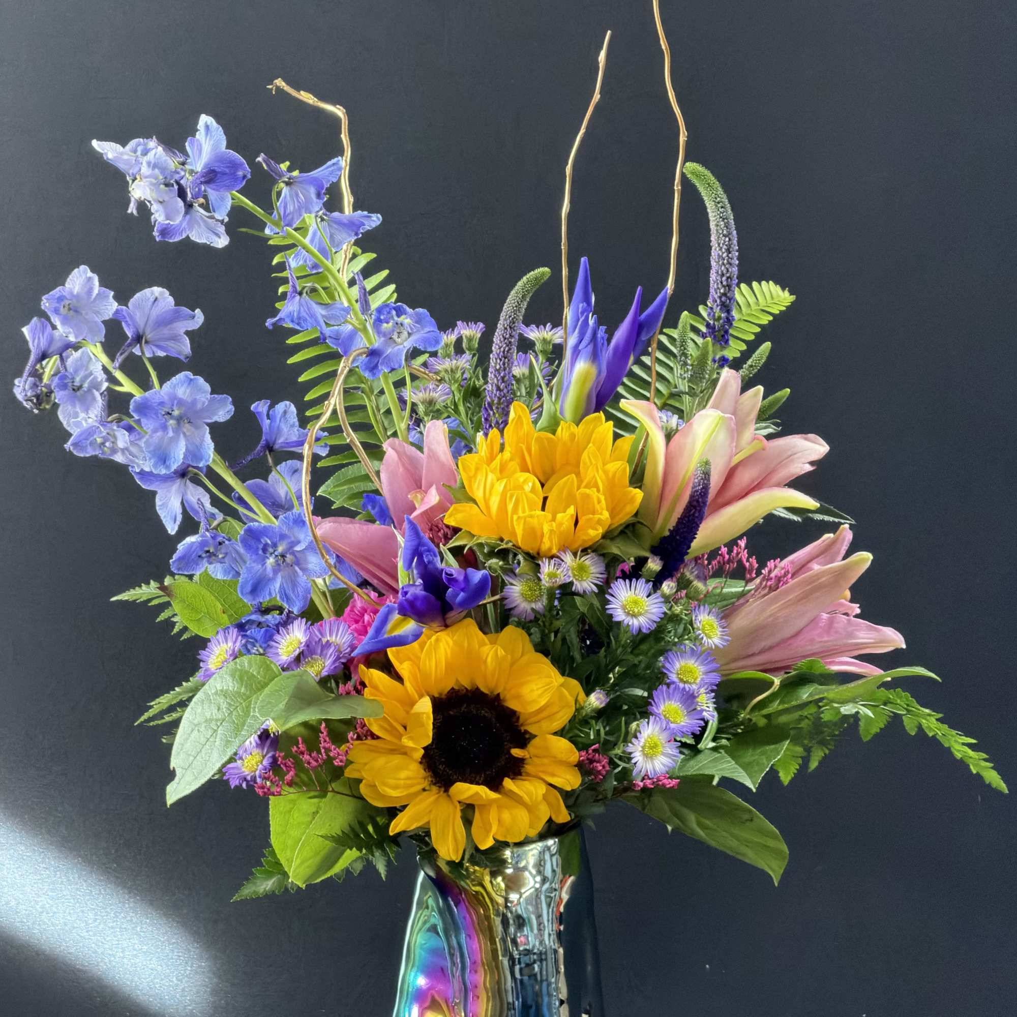 Tall mixed arrangement with sunflowers, pink lilies, and blue blooms in an iridescent vase