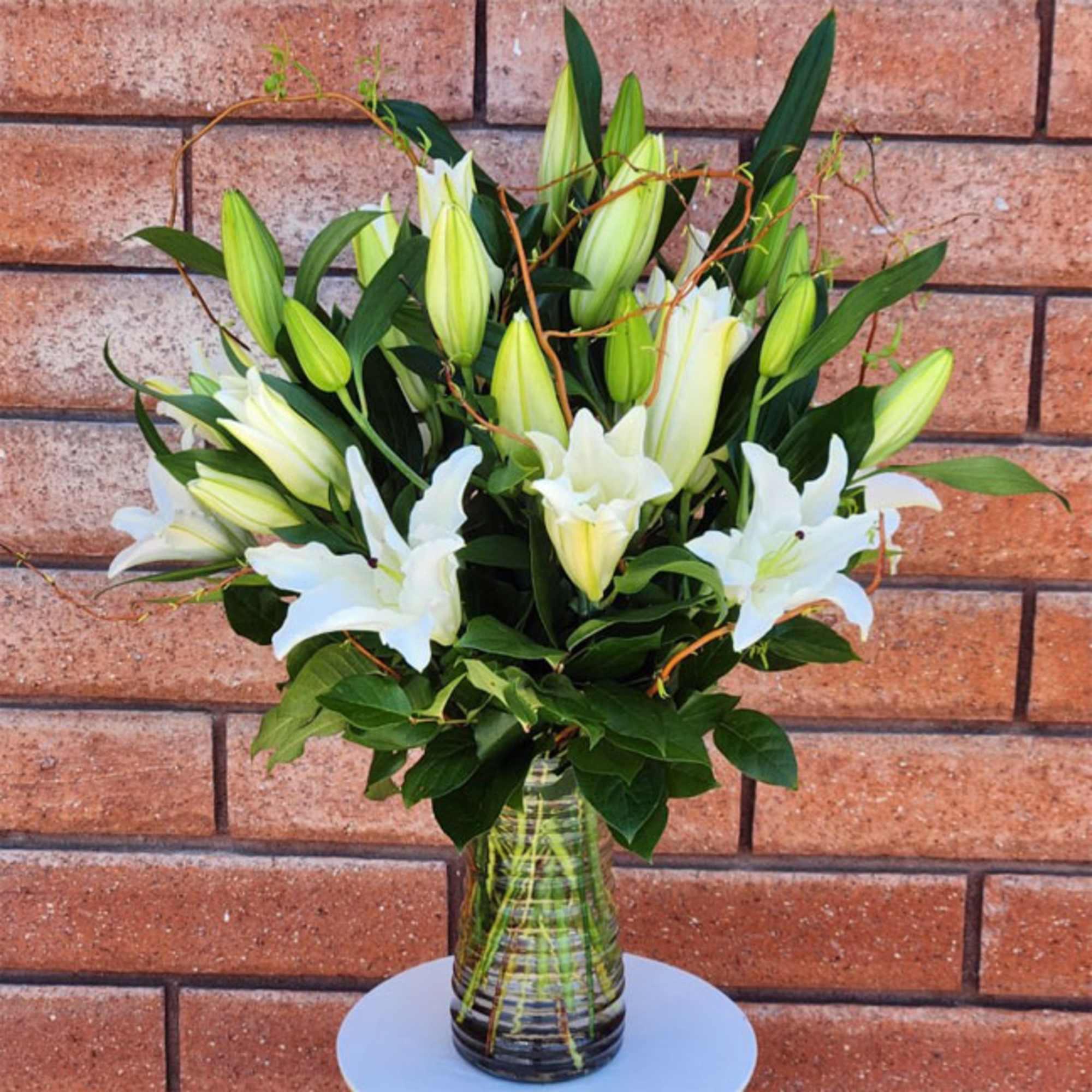 Tall arrangement of white lilies in a clear glass vase against a brick wall