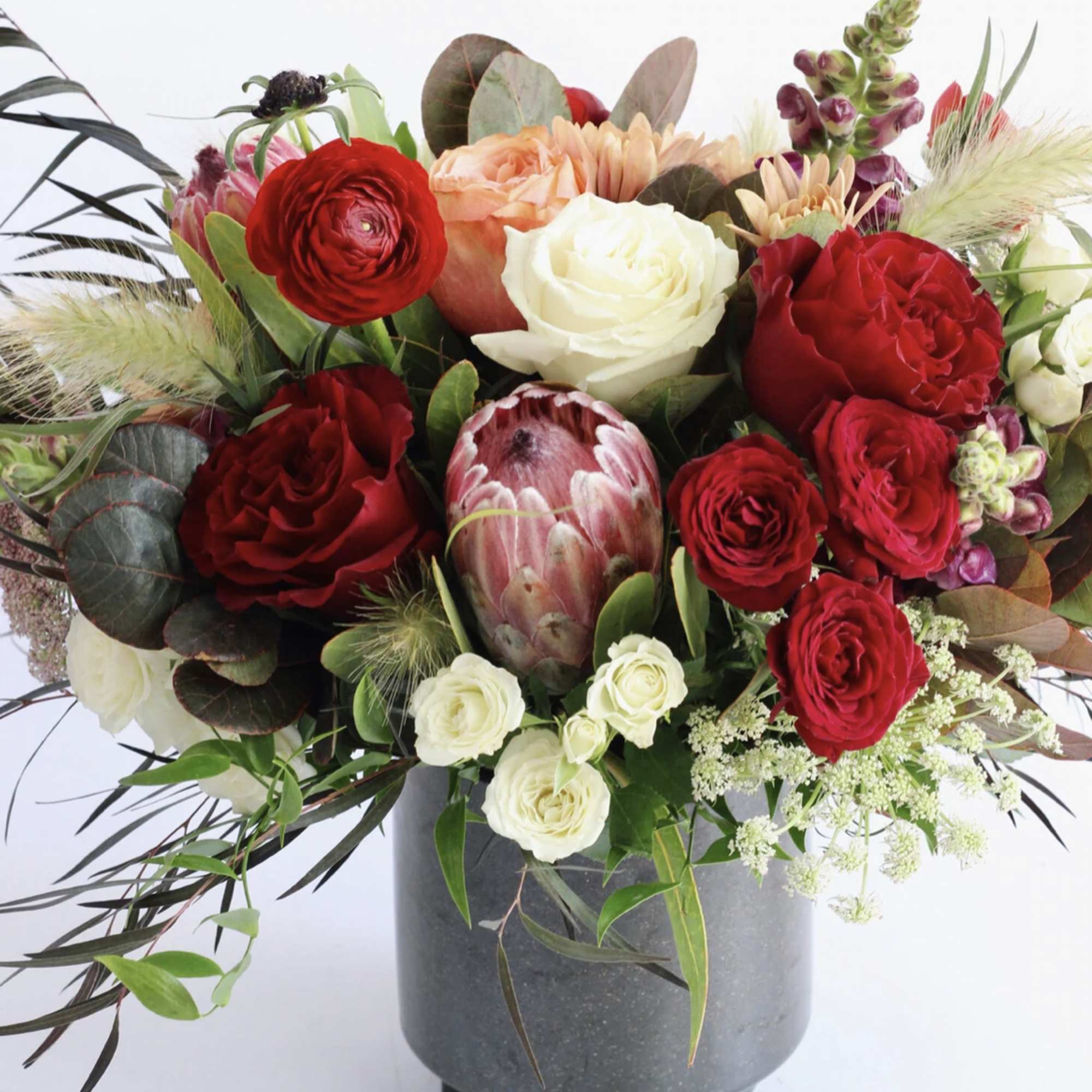 Lush arrangement of red and cream roses, protea, and mixed blooms in a dark gray vase