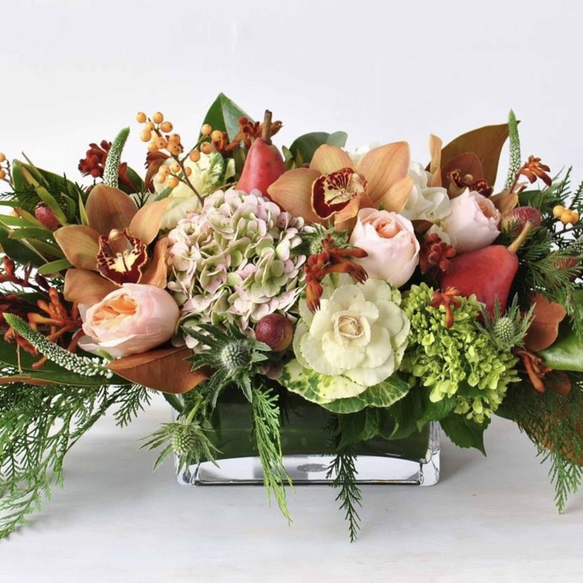 Low rectangular centerpiece with peach roses, orchids, hydrangea, berries, and evergreen accents in a glass vase