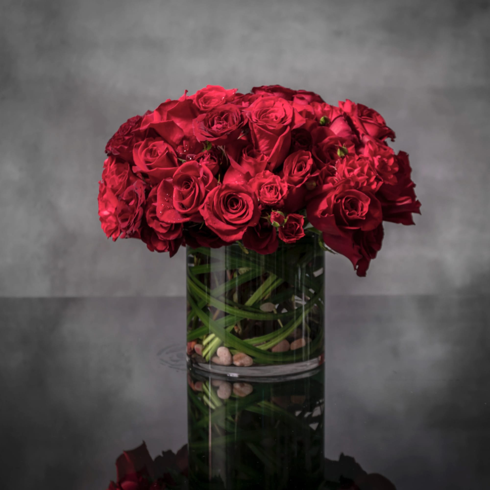 Low cylinder vase of clustered red roses with decorative green stems and pebbles at the base.