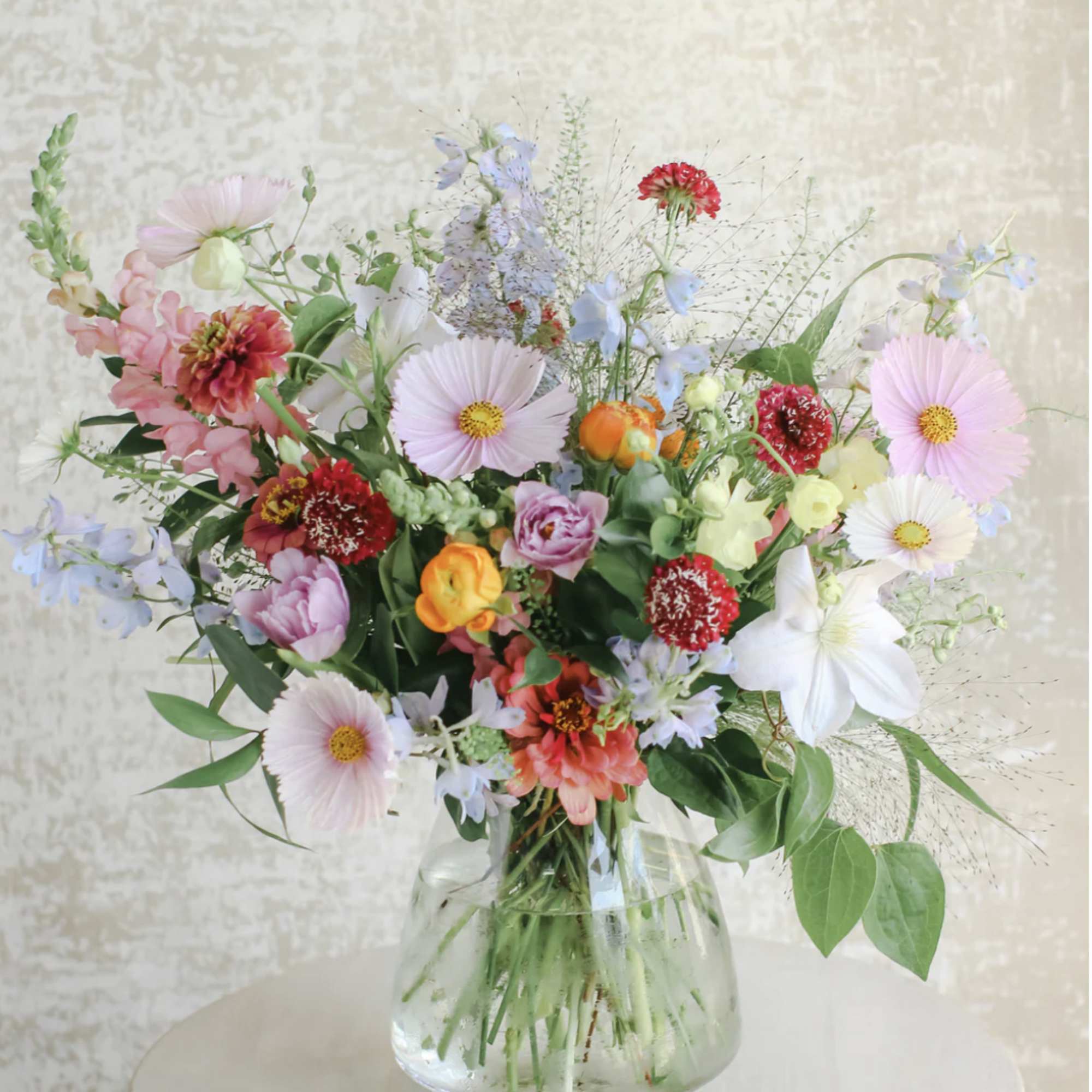Loose garden-style bouquet of mixed pastel and bright flowers in a clear glass vase.