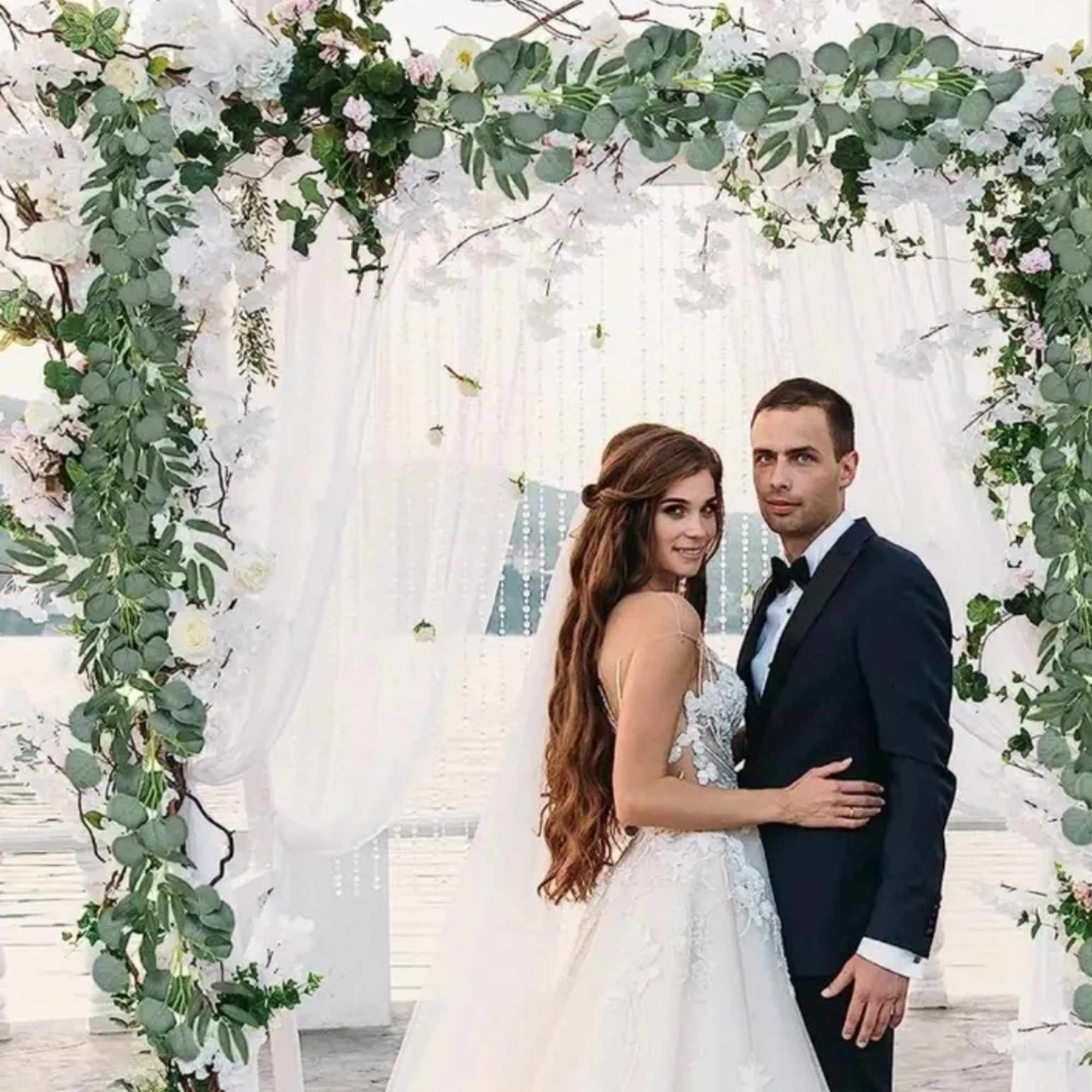 Wedding couple standing under a white floral and greenery arch with draped fabric backdrop