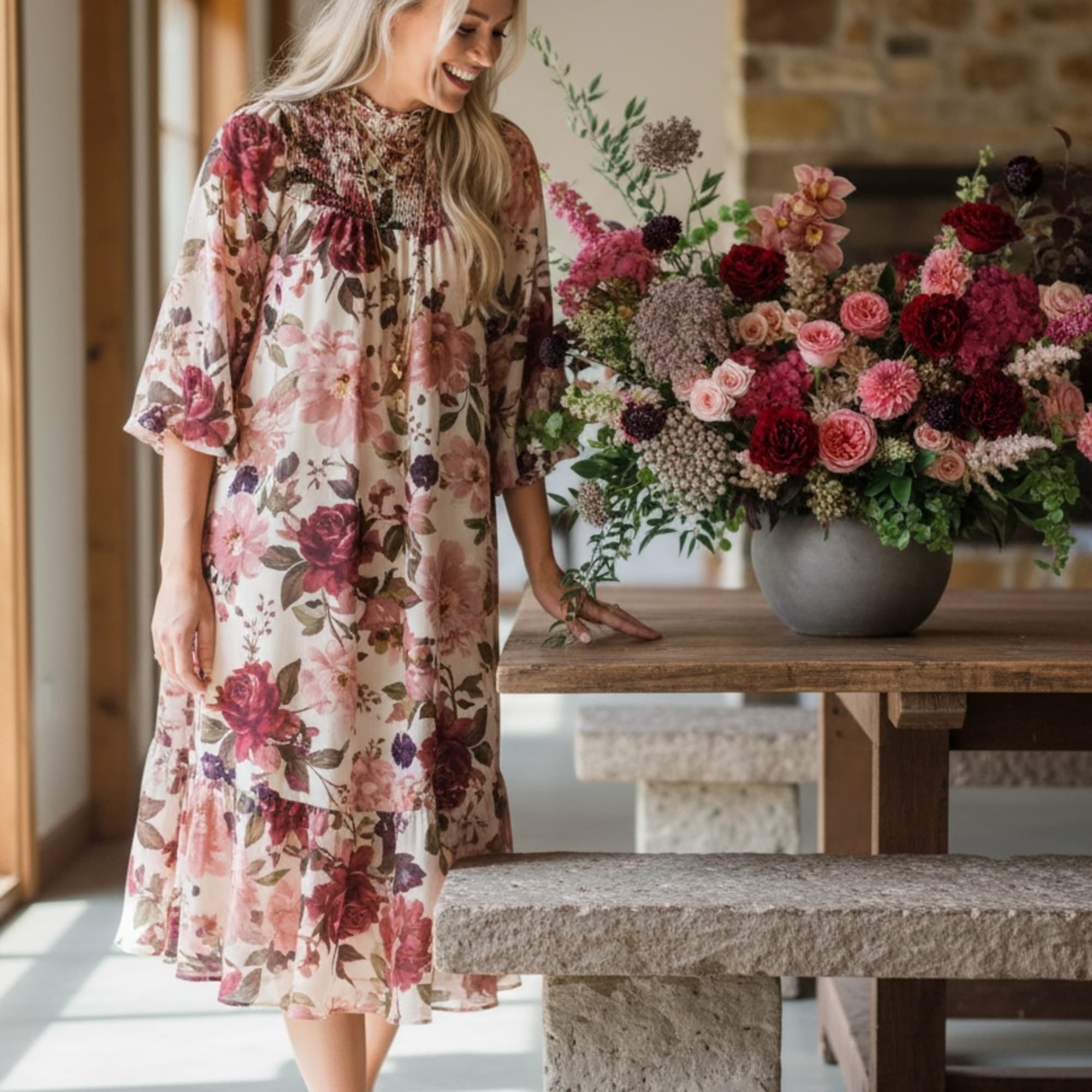 Large pink and burgundy flower arrangement in a gray bowl on a wooden table beside a smiling woman.