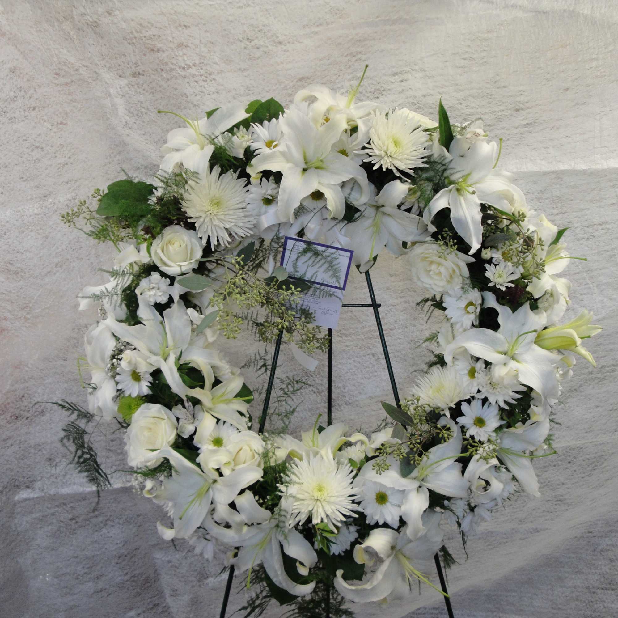 White floral wreath on a black stand with lilies and daisies