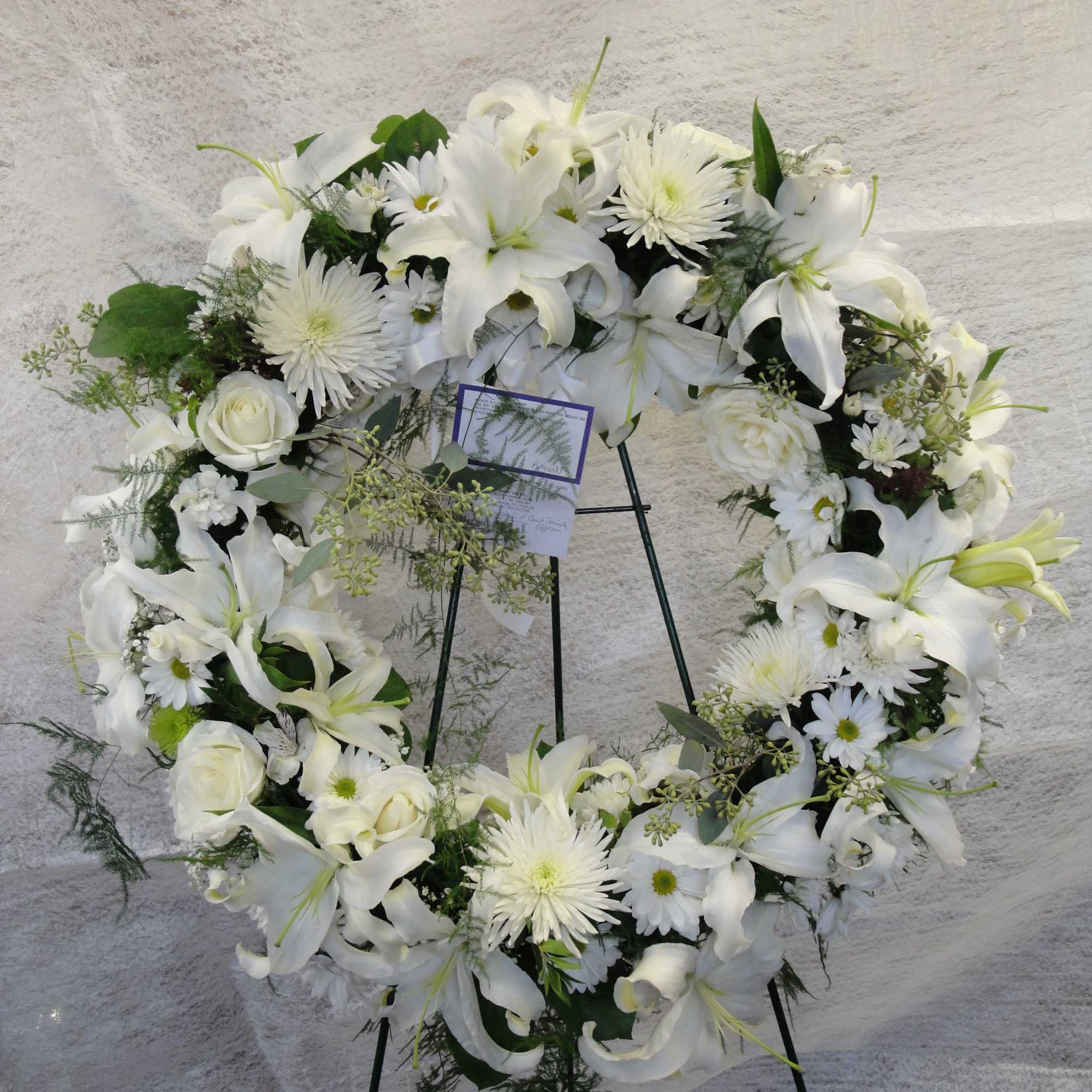White floral wreath on a black stand with lilies, roses, and daisies