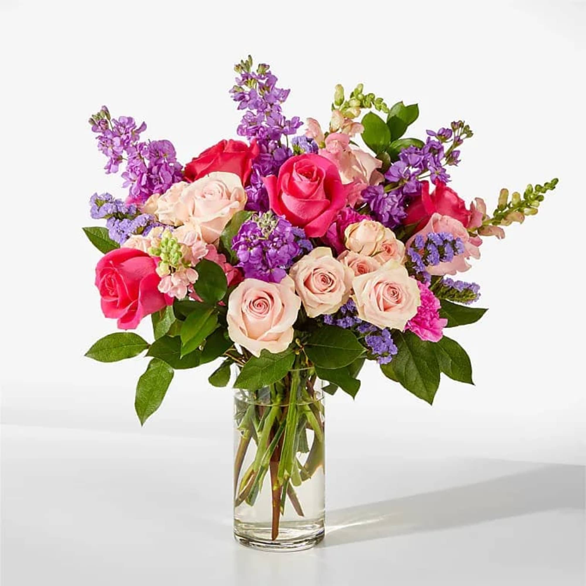Bouquet of hot pink and blush roses with purple flowers arranged in a clear glass vase.