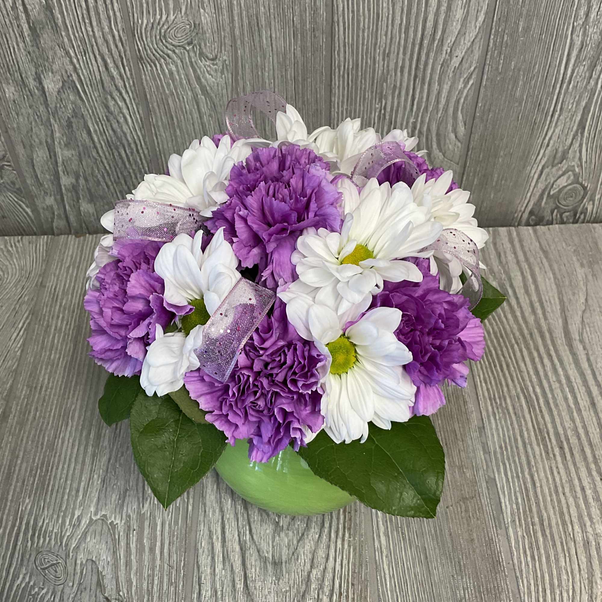 Purple carnations and white daisies in a green vase with ribbon