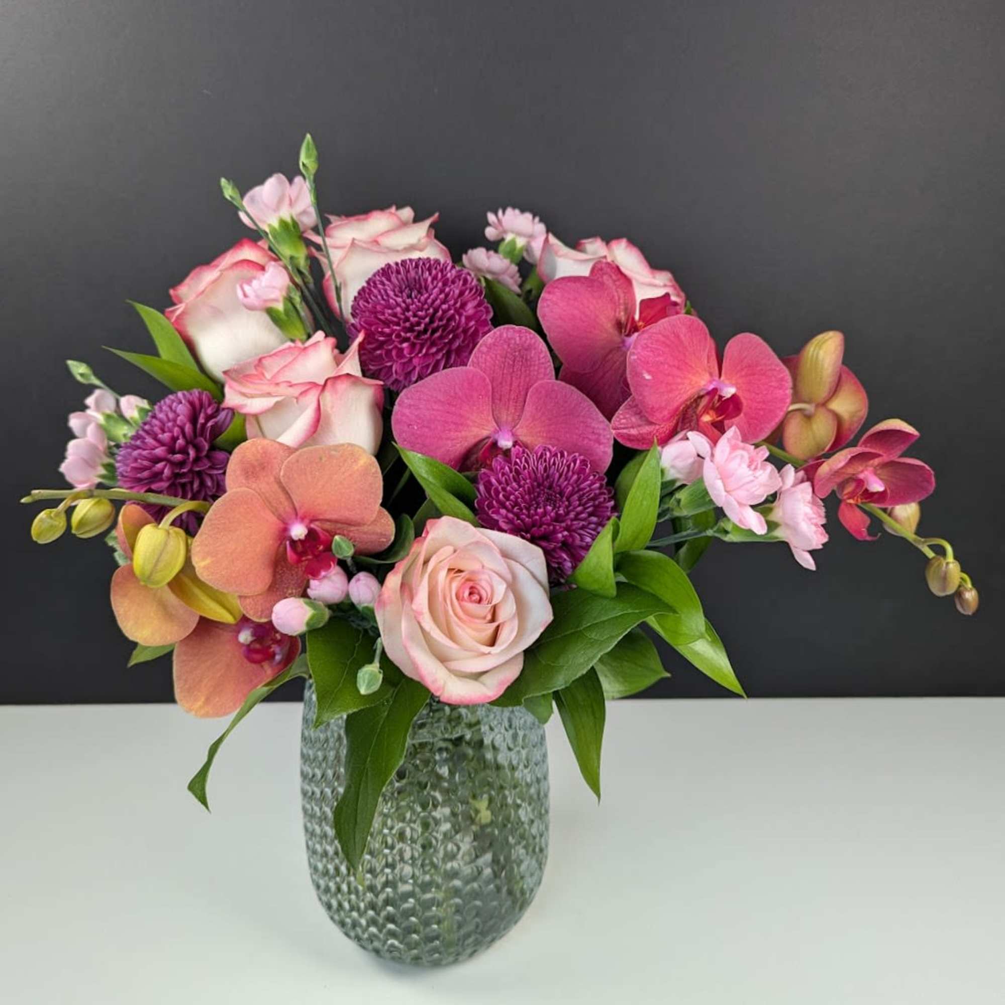 Pink and purple arrangement of roses, orchids, and mums in a textured glass vase