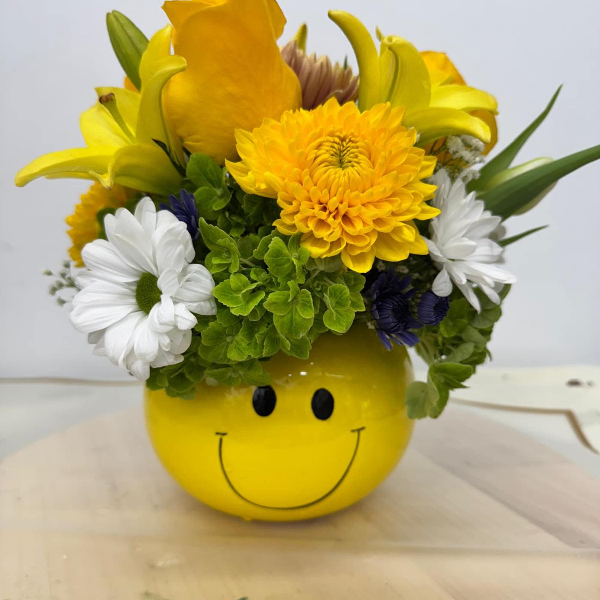 Yellow smiley-face container holding a mixed bouquet of yellow, white, and purple flowers