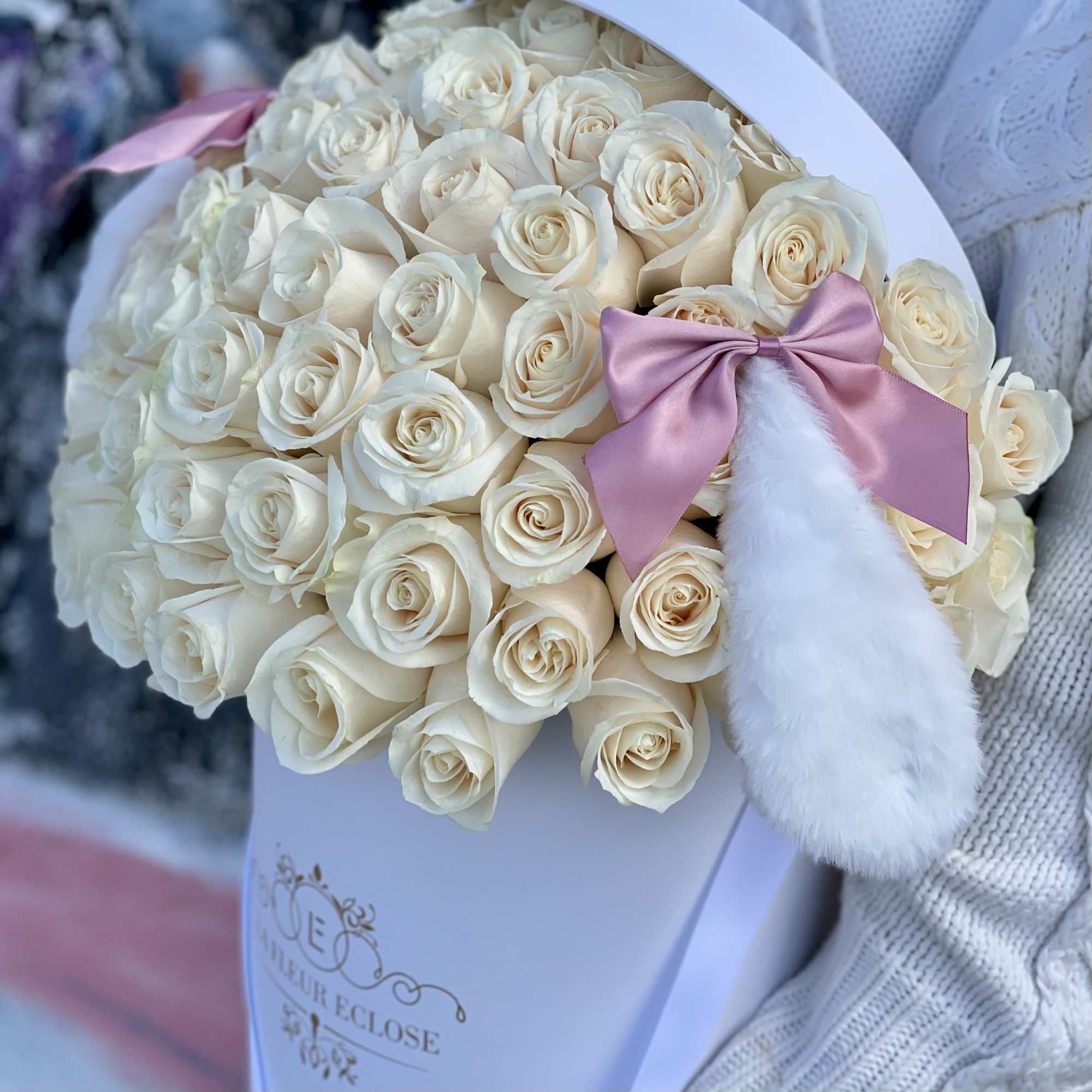 Round white box filled with cream roses, trimmed with pink bows and a white faux fur accent.