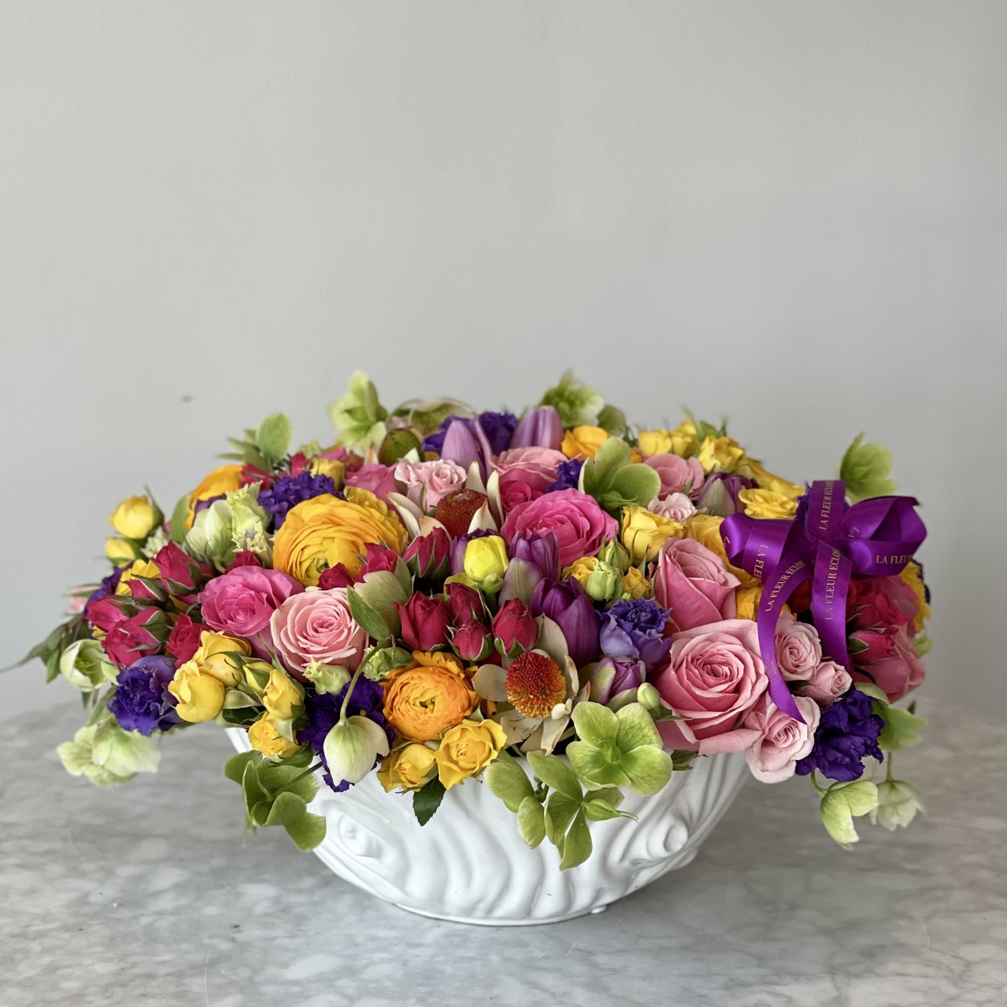 Mixed bouquet of pink, yellow, purple, and green flowers in a white bowl vase