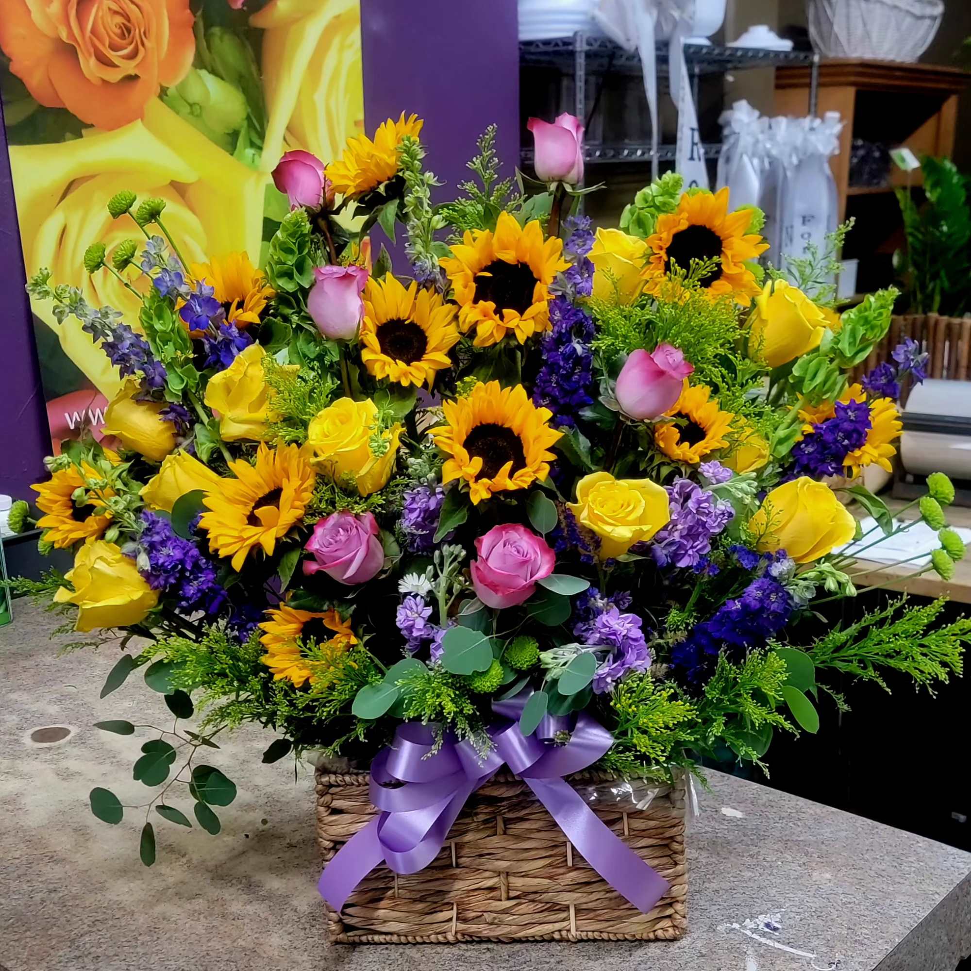 Large basket arrangement with sunflowers, yellow and pink roses, and purple flowers tied with a lavender bow.