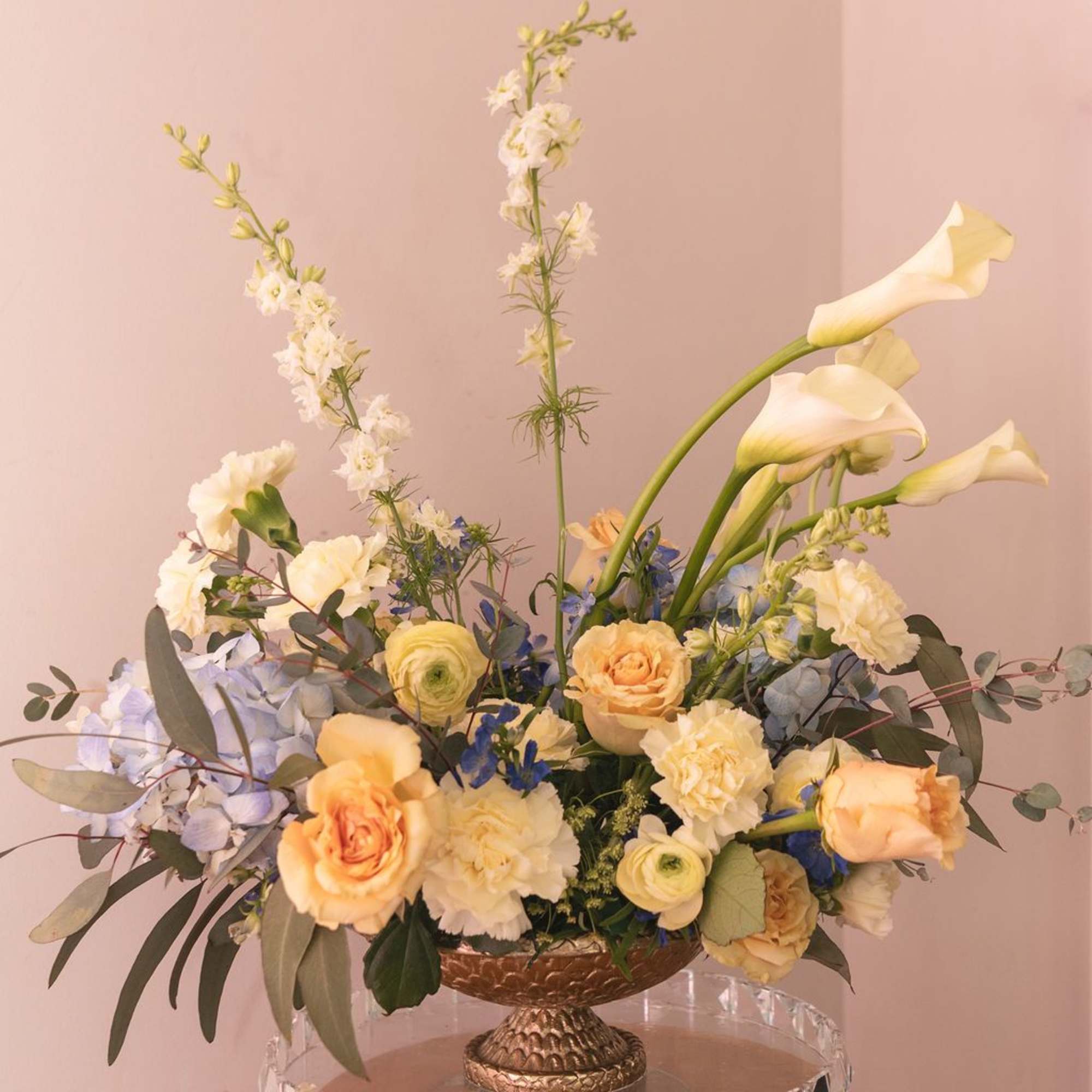 Pastel floral arrangement with calla lilies, roses, and blue flowers in a pedestal bowl