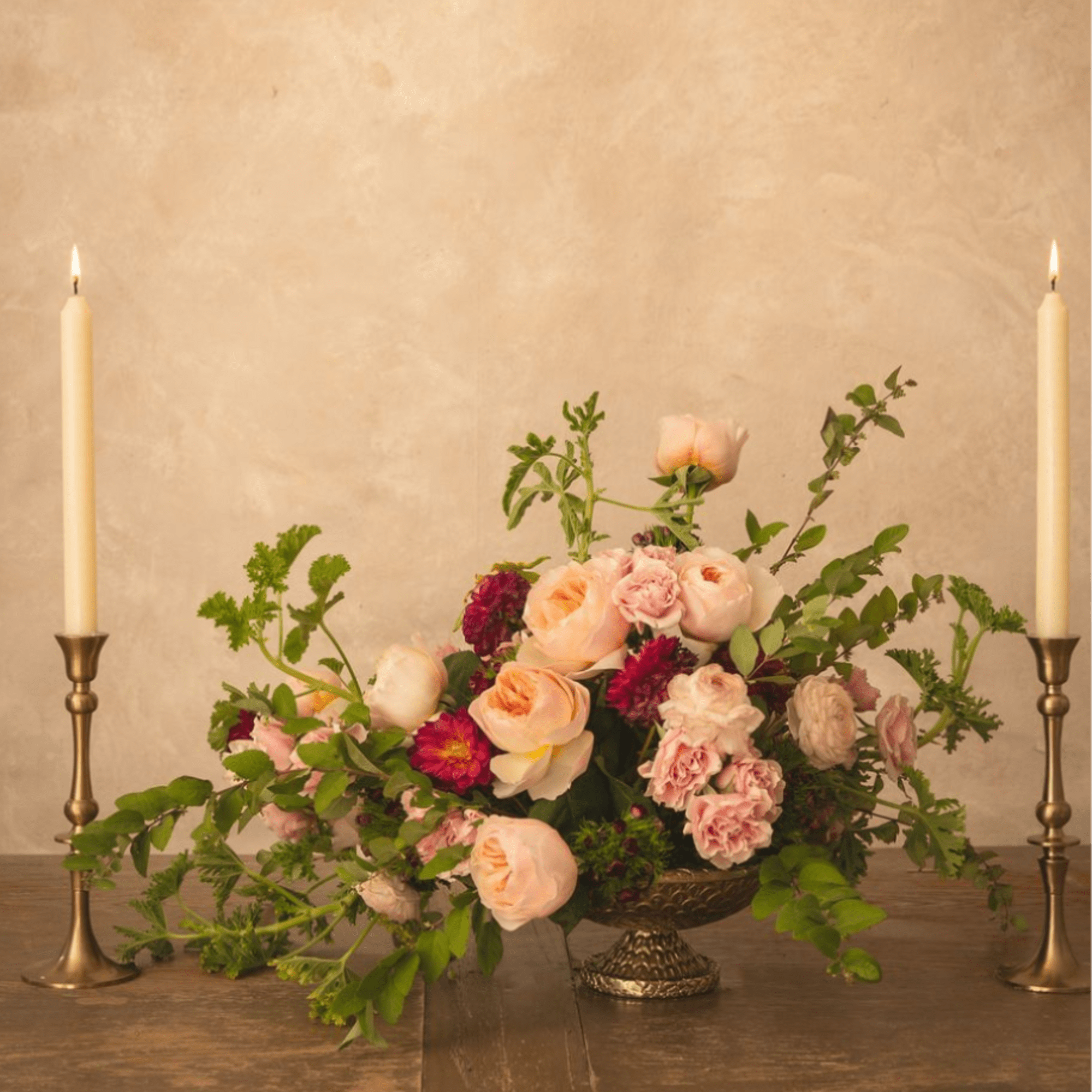 Low floral centerpiece with blush and peach roses on a pedestal, flanked by two lit taper candles