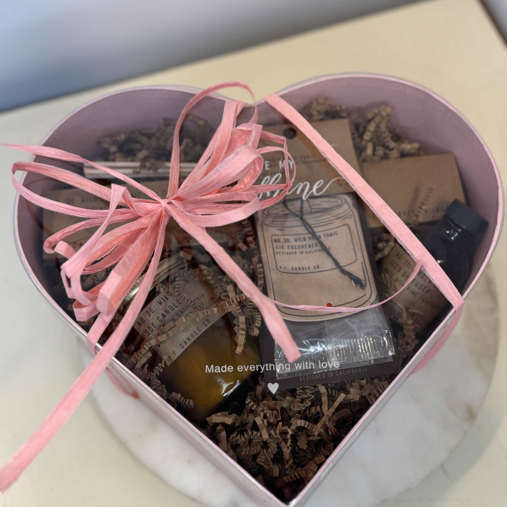 Heart-shaped gift box with candles and small packaged items tied with a pink ribbon