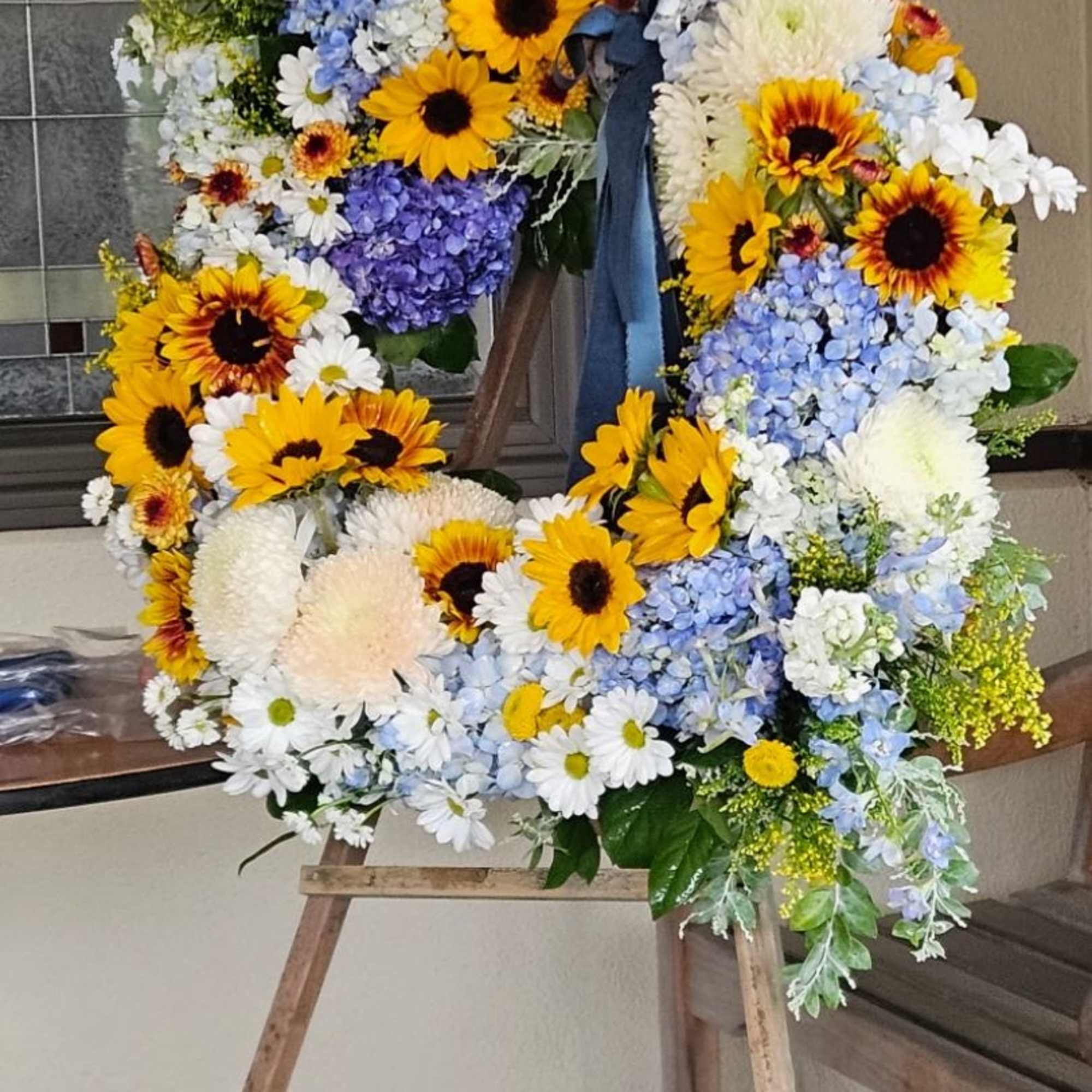 Large floral wreath with sunflowers, blue hydrangeas, and white daisies on an easel