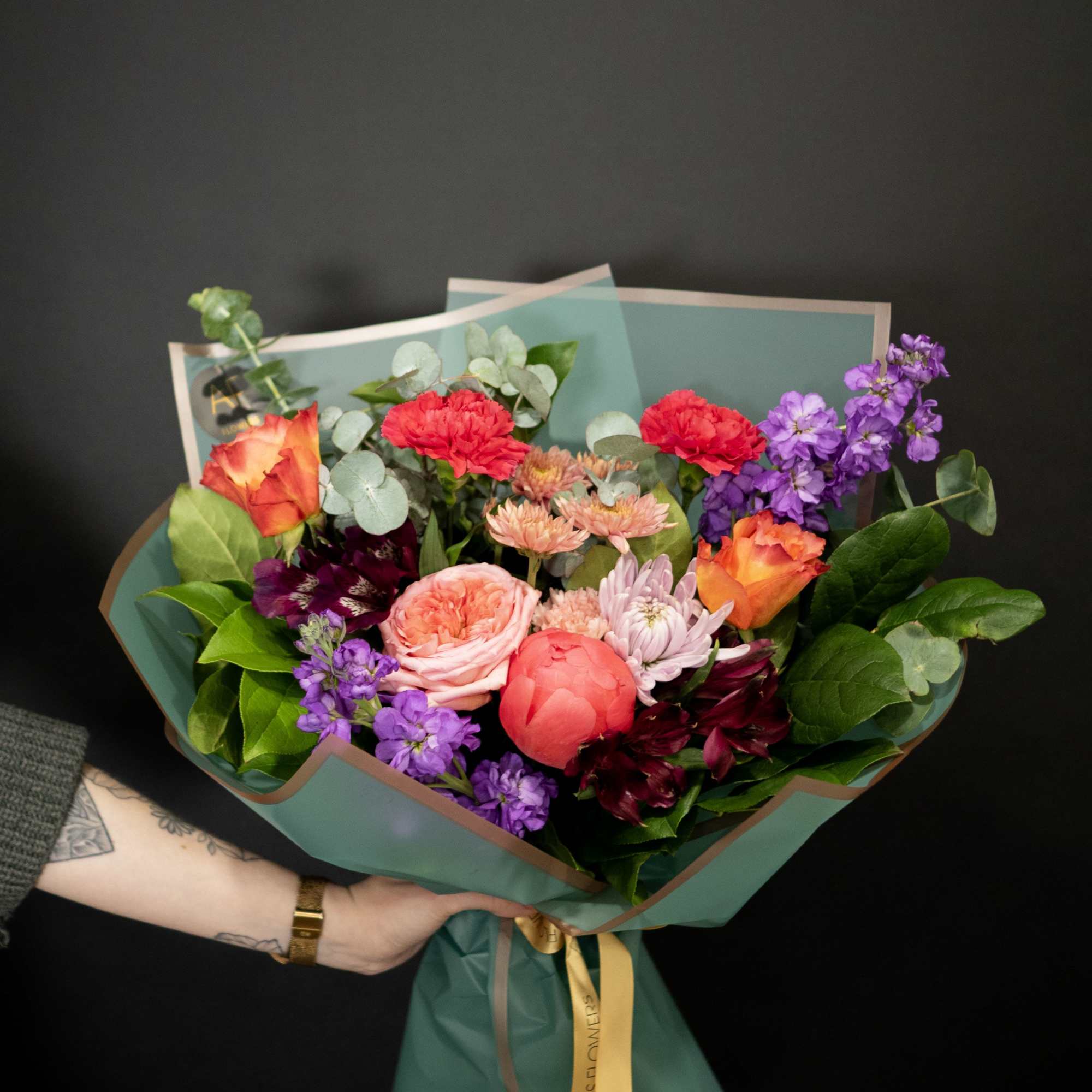 Mixed bouquet of roses, carnations, and purple blooms wrapped in teal paper