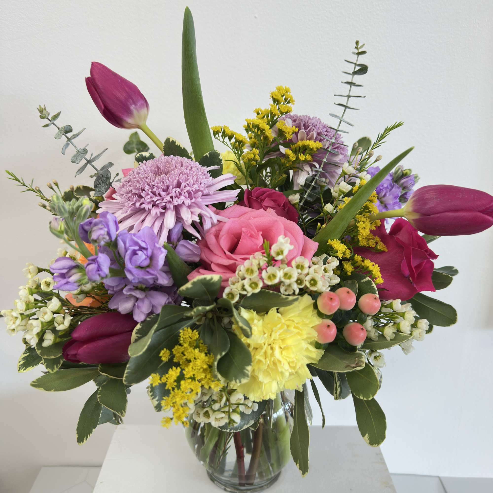 Bright mixed bouquet with pink roses, purple mums, yellow carnations and tulips in a clear glass vase