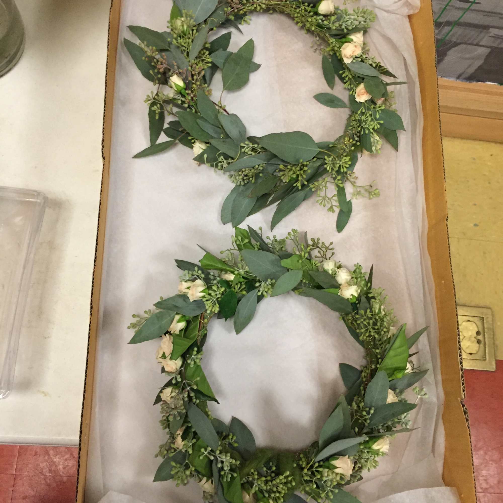Two floral wreaths with white blooms and greenery in a box